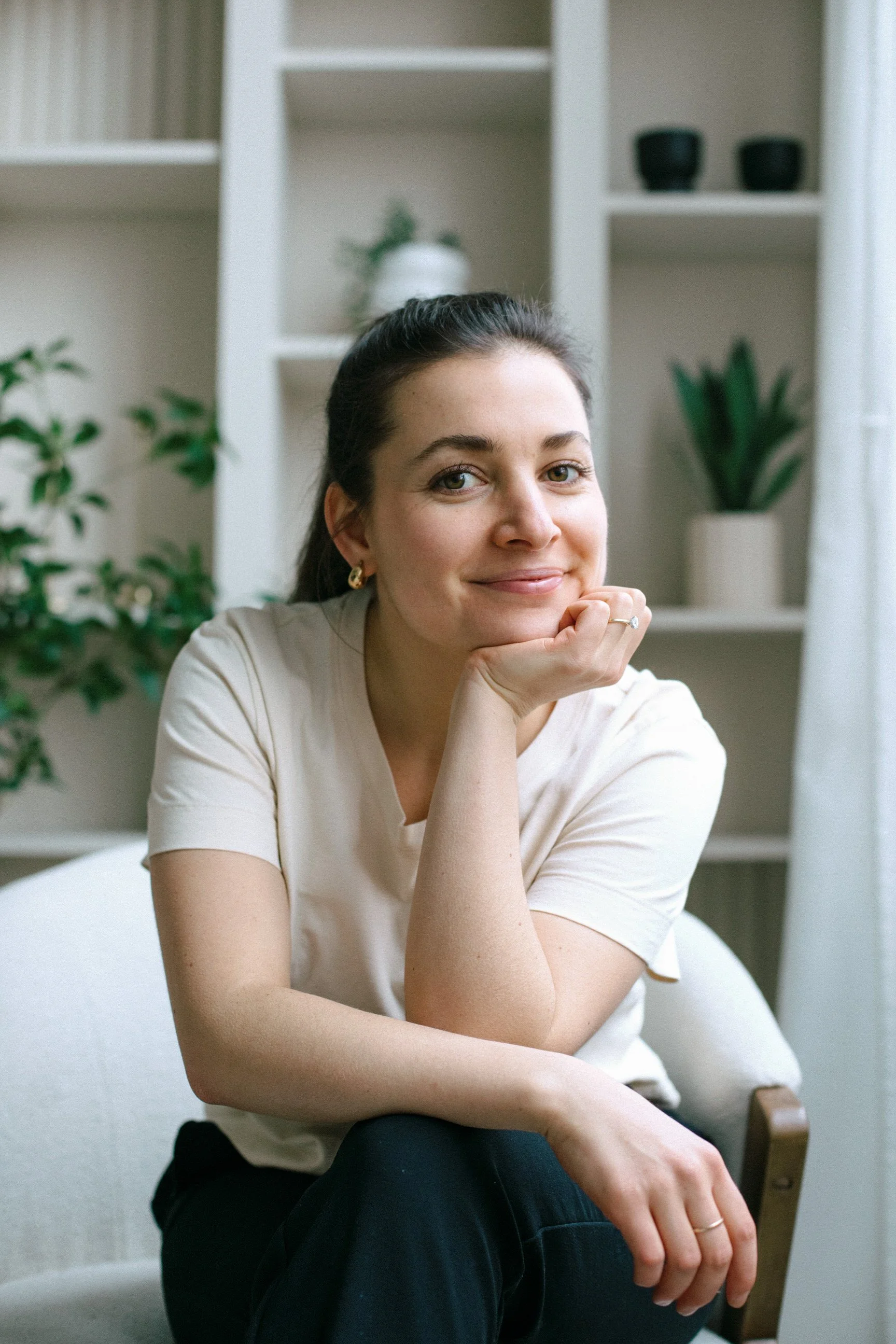 A young woman with dark hair, wearing a light-colored t-shirt and gold earrings, sitting in a room with white shelves and green plants, smiling gently and resting her chin on her hand.