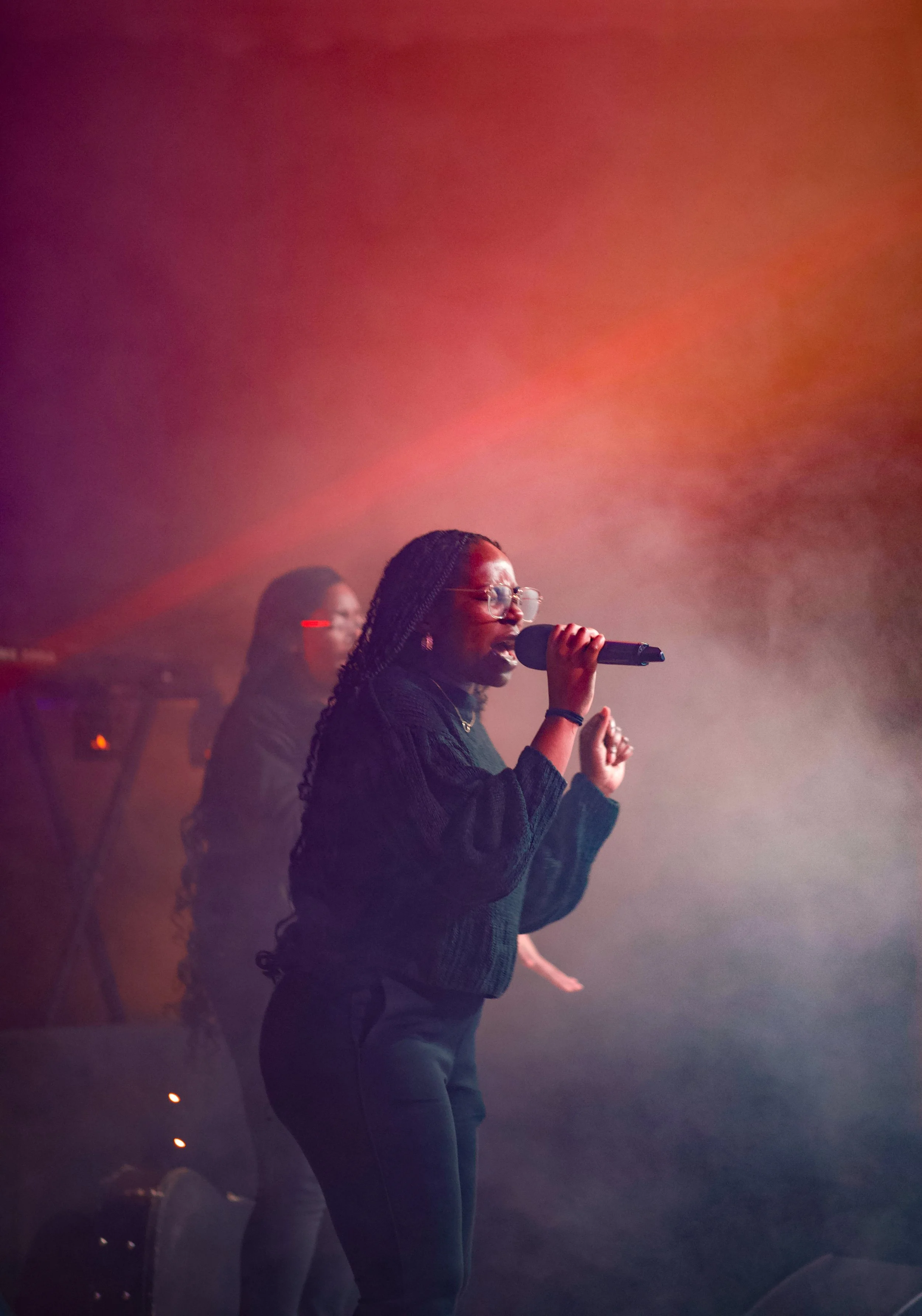 A woman wearing glasses and a braided hairstyle singing into a microphone in a smoky, colorful lighted concert or performance setting.