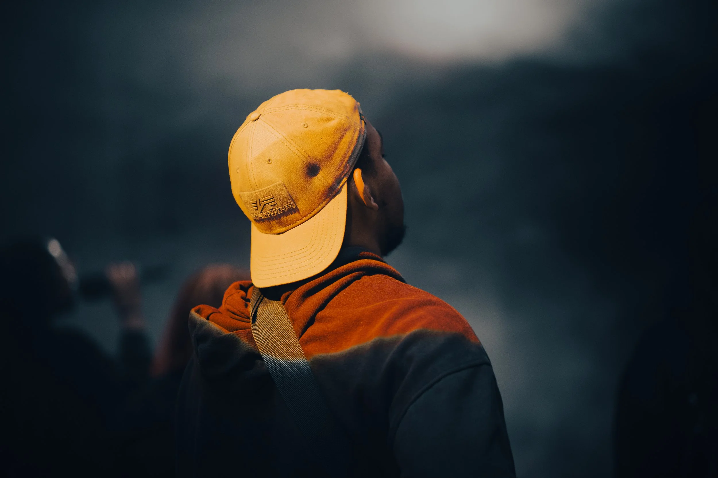 A person wearing a yellow cap and orange hoodie, looking out into a dark, cloudy sky.