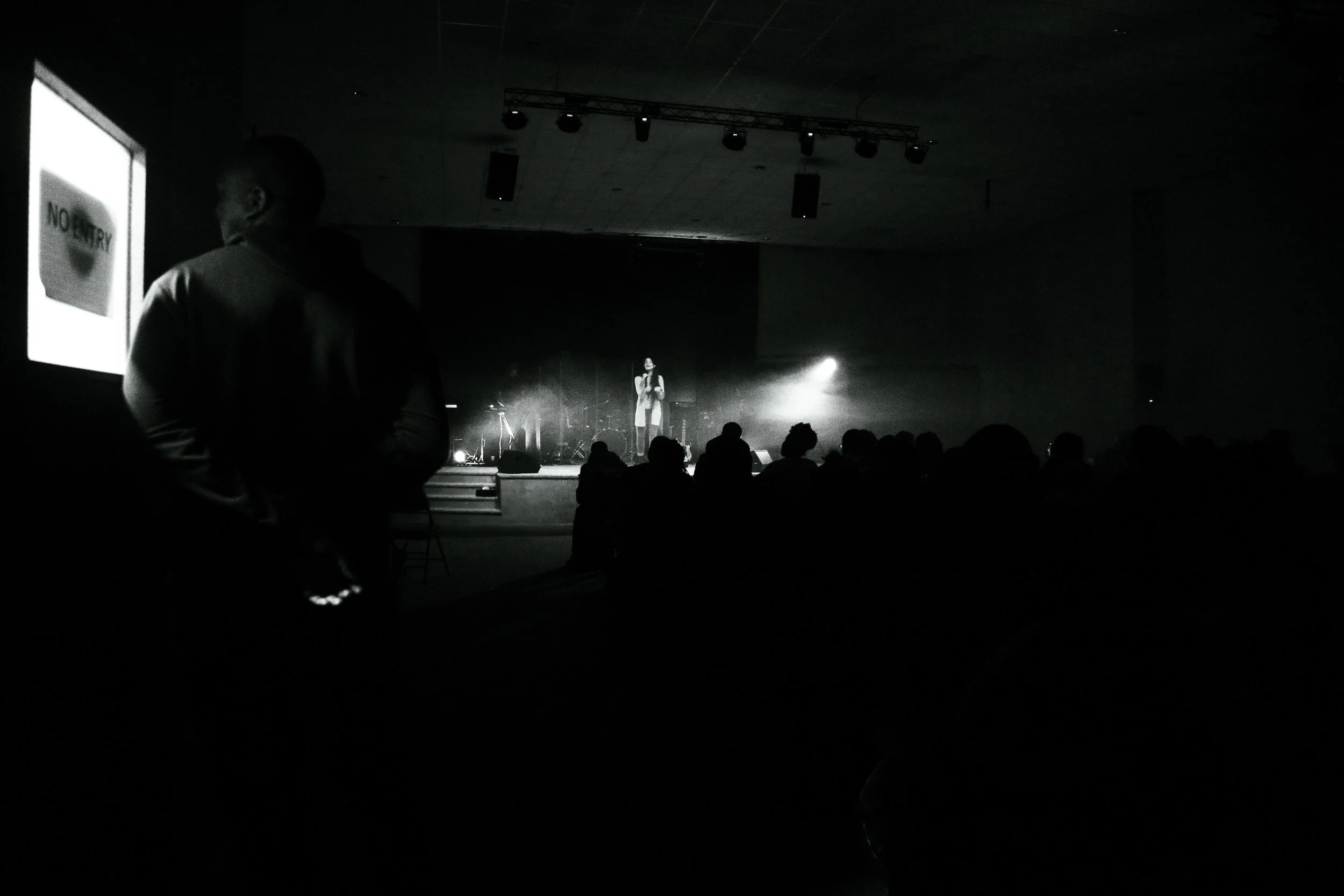 A person standing on stage in front of an audience, with stage lights and equipment, while audience members are seated facing the stage. The setting appears to be an indoor auditorium or conference space.