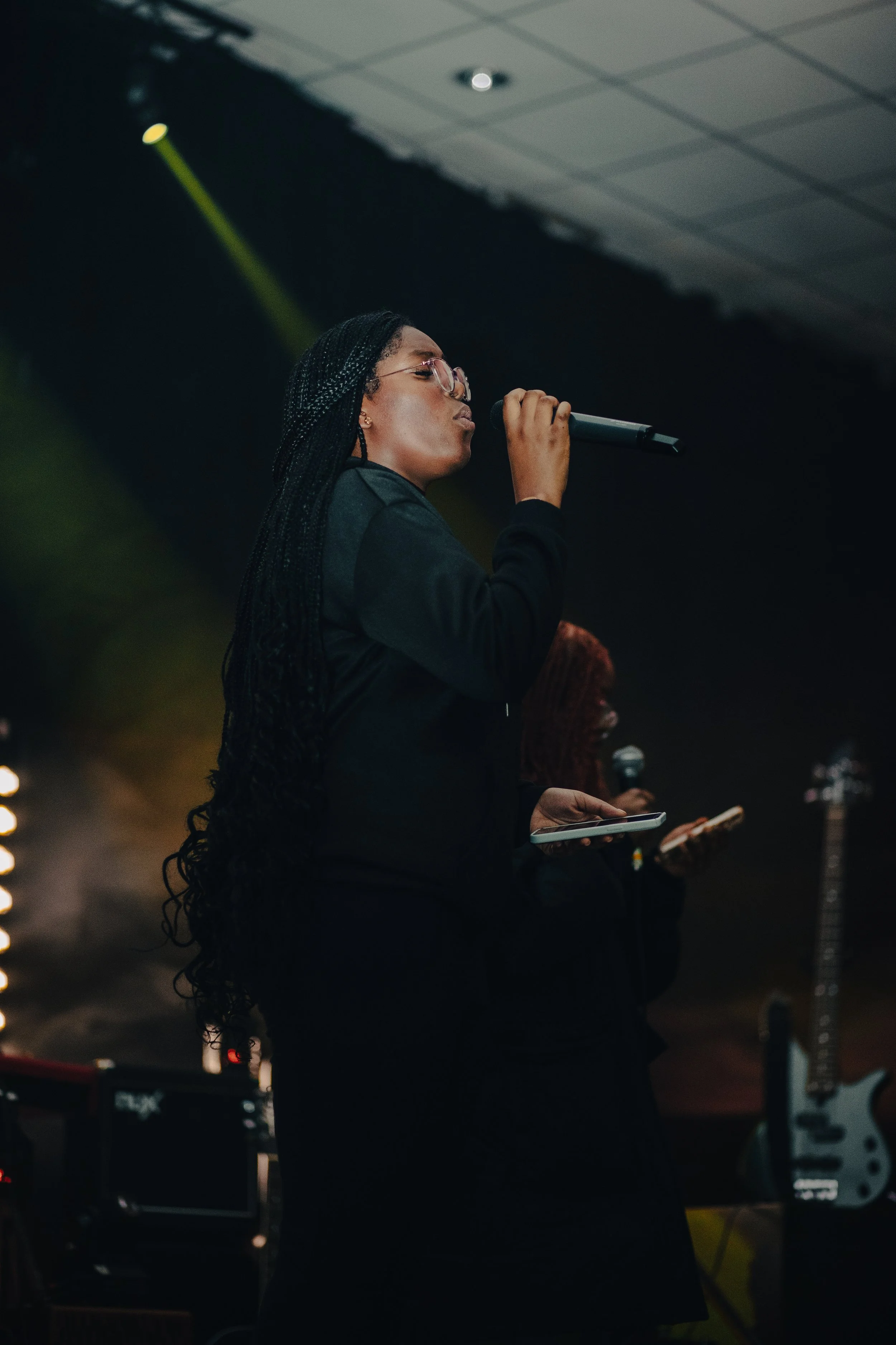 A woman with long braided hair and glasses singing into a microphone on stage, holding a tablet, with musical equipment and another person in the background.