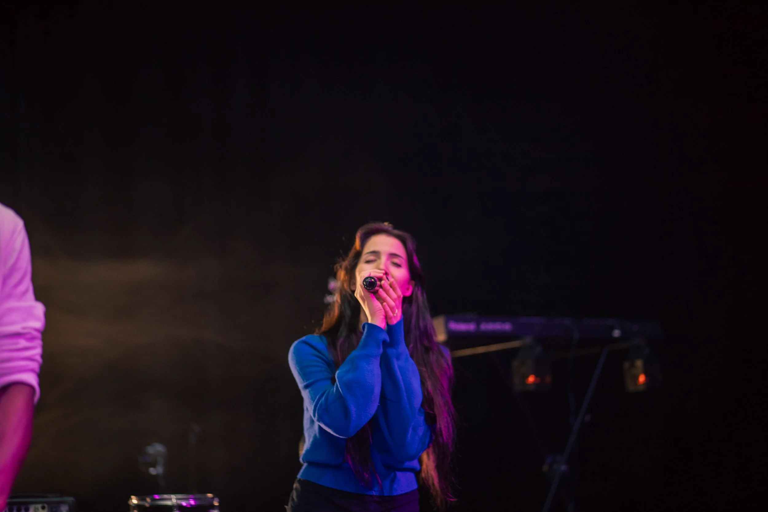 Young woman singing into a microphone on stage with dark background and musical equipment.