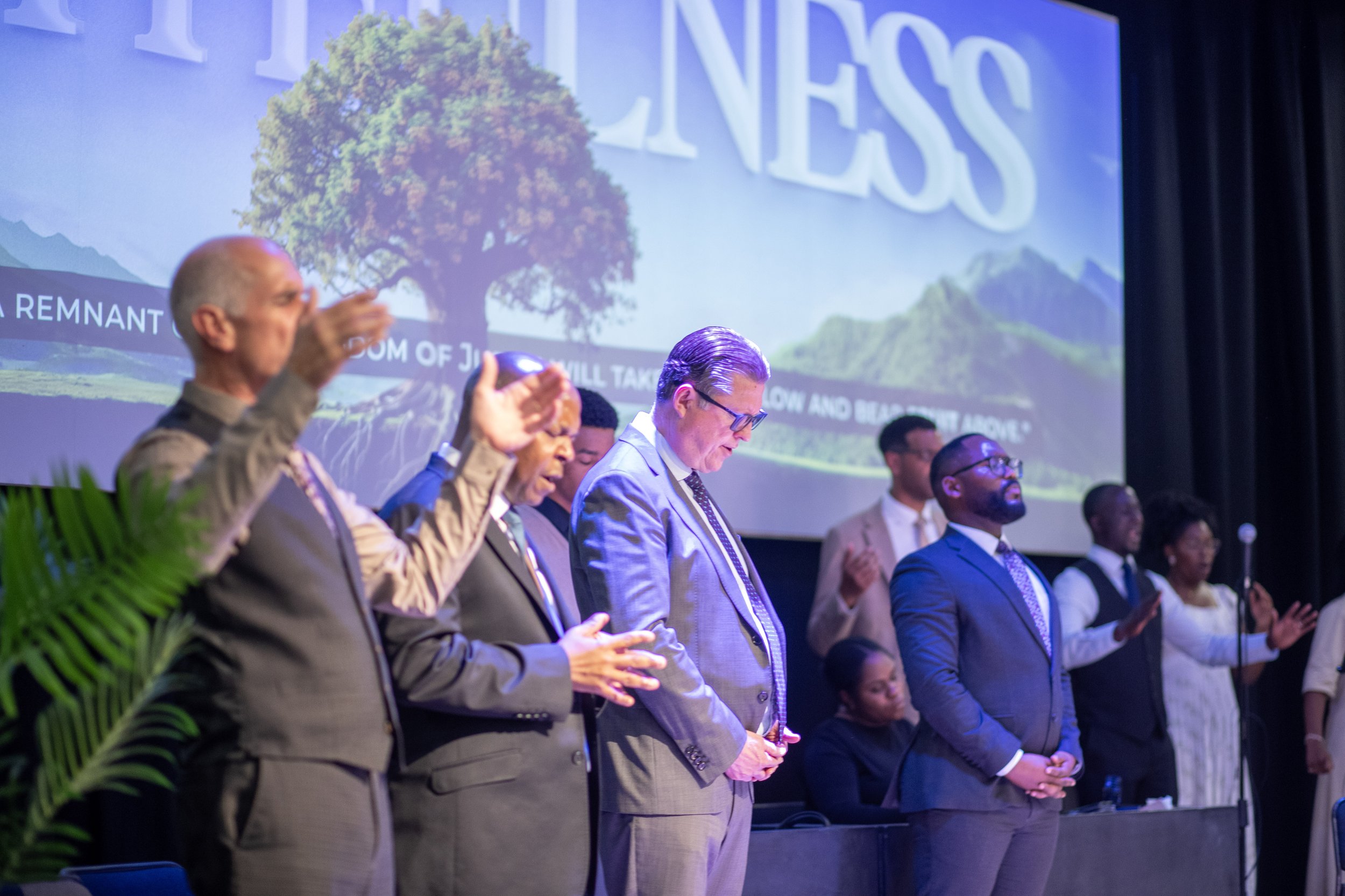 A group of diverse people, mostly men, standing in a row on a stage with their eyes closed and hands raised, participating in an event. Behind them, a large screen displays a tree, mountains, and the word 'KINDNESS' in large white letters.