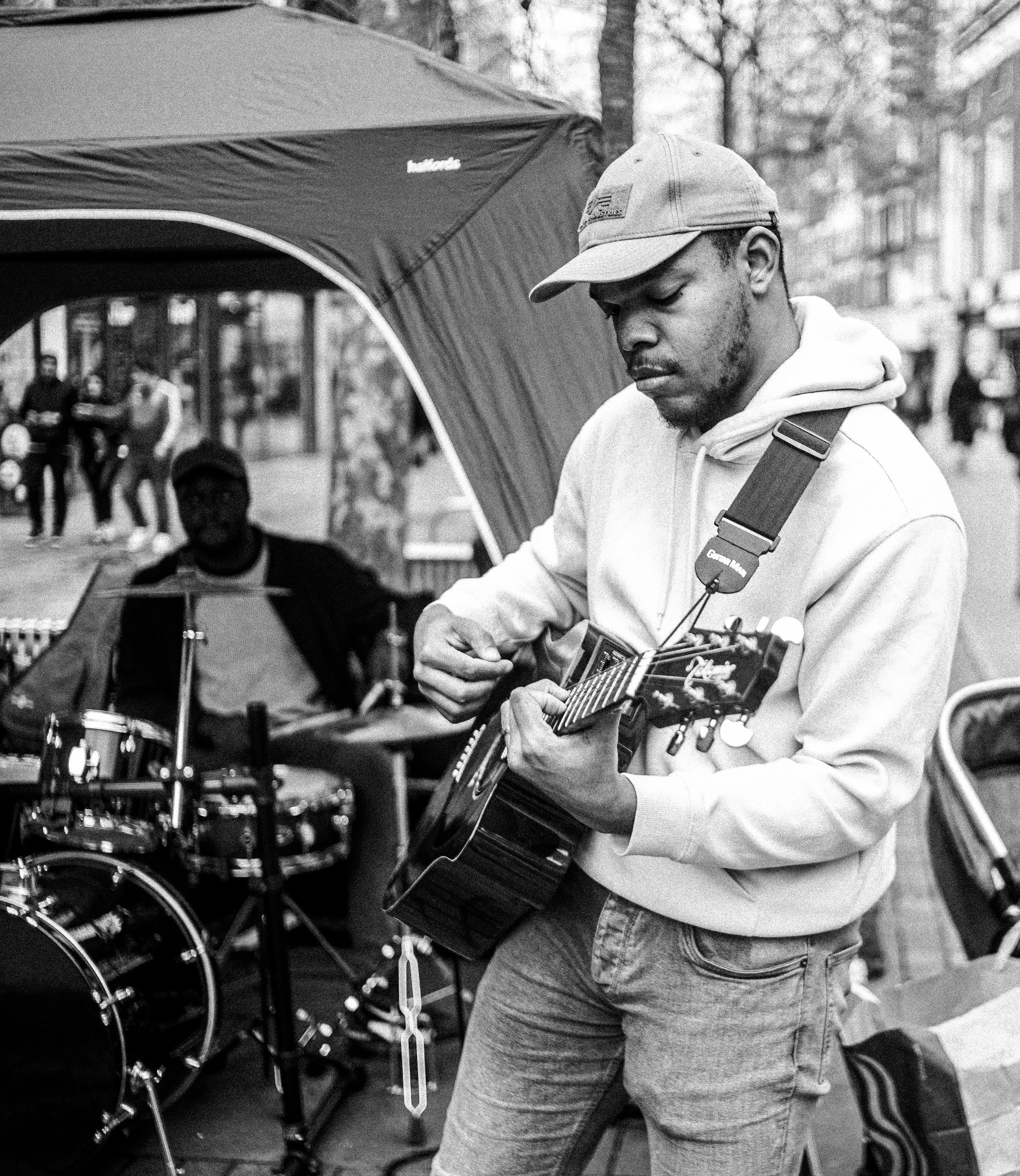 A man in a hoodie and cap playing an acoustic guitar outdoors near a row of drum sets, with a few people blurred in the background.