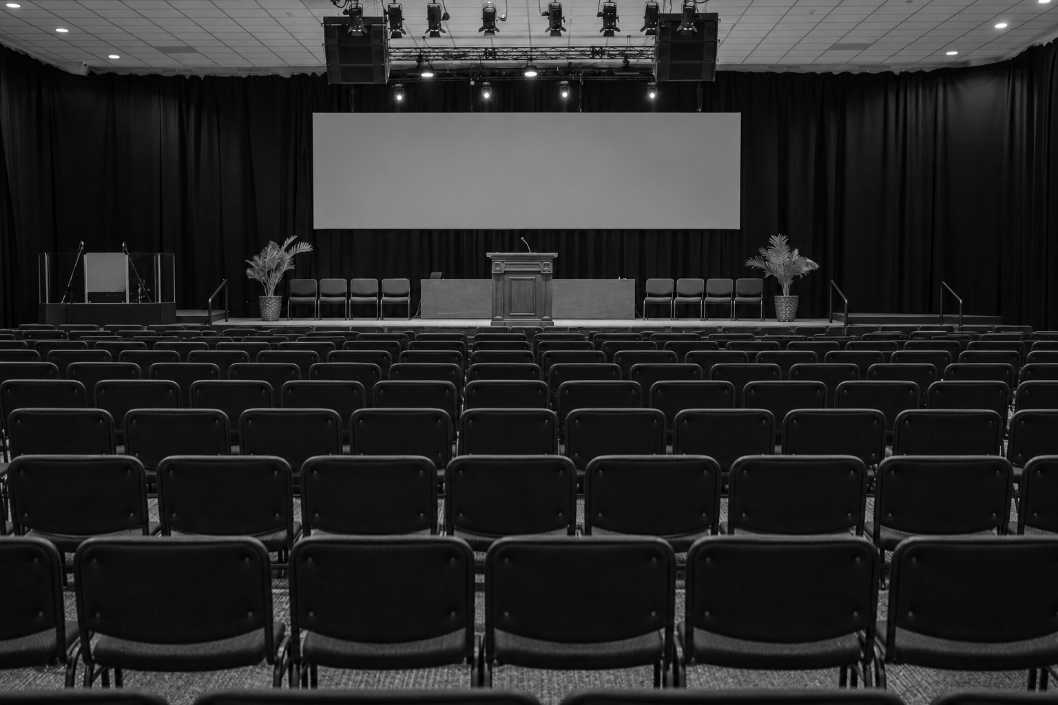 Empty auditorium with rows of black chairs facing a stage with a wooden podium, potted plants, and a large blank screen.