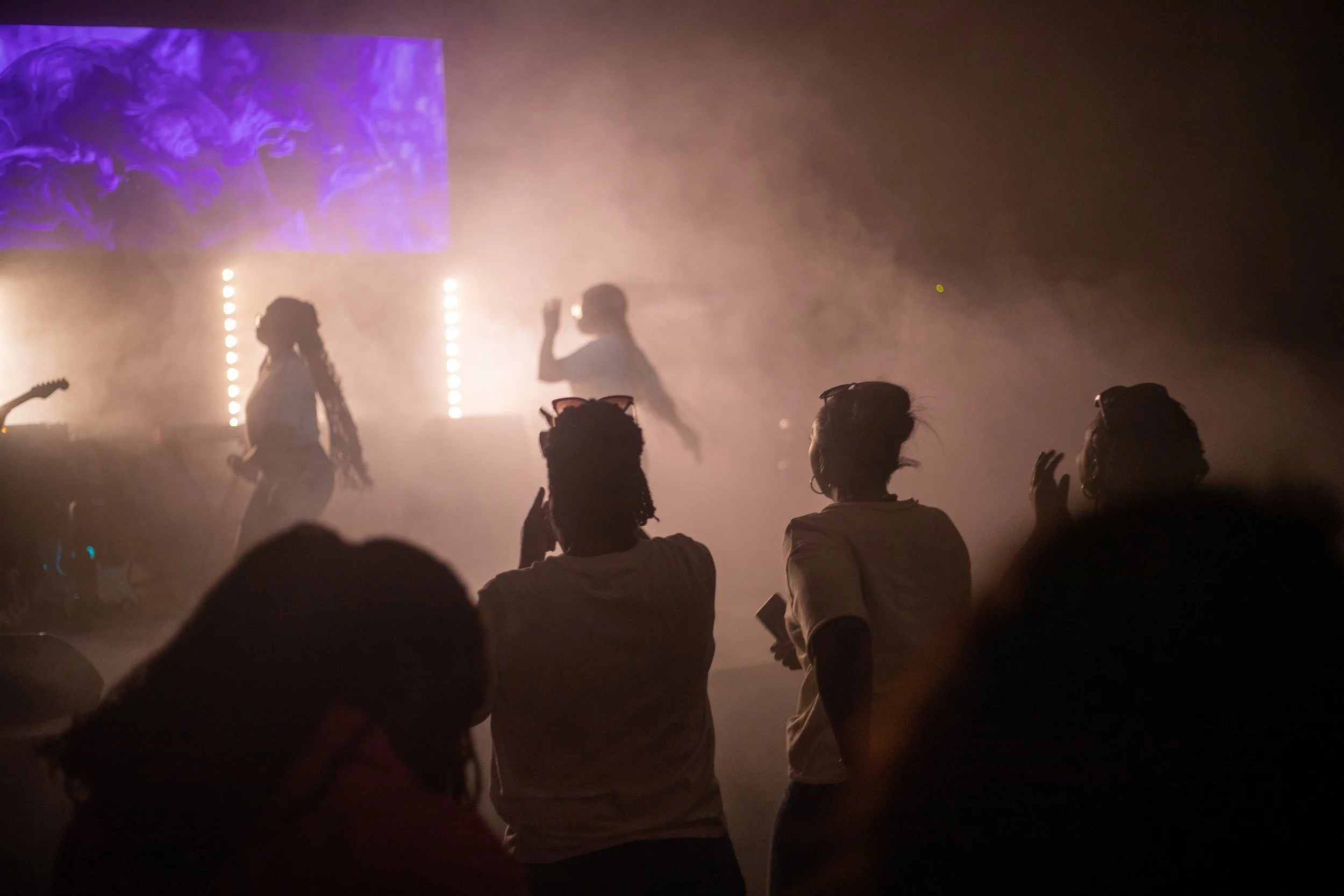 People at a concert or event, silhouetted against a bright, foggy background with purple and white lighting, some dancing and raising hands.