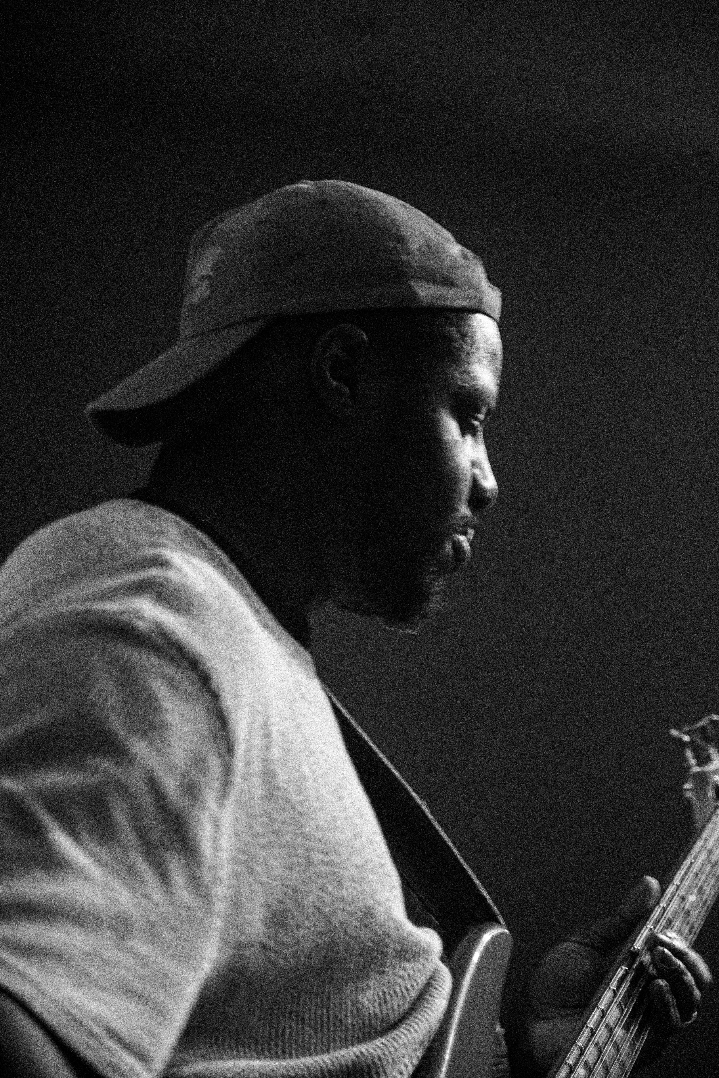 A man wearing a baseball cap and a sweatshirt is playing an electric guitar, viewed in profile, with a dark background.