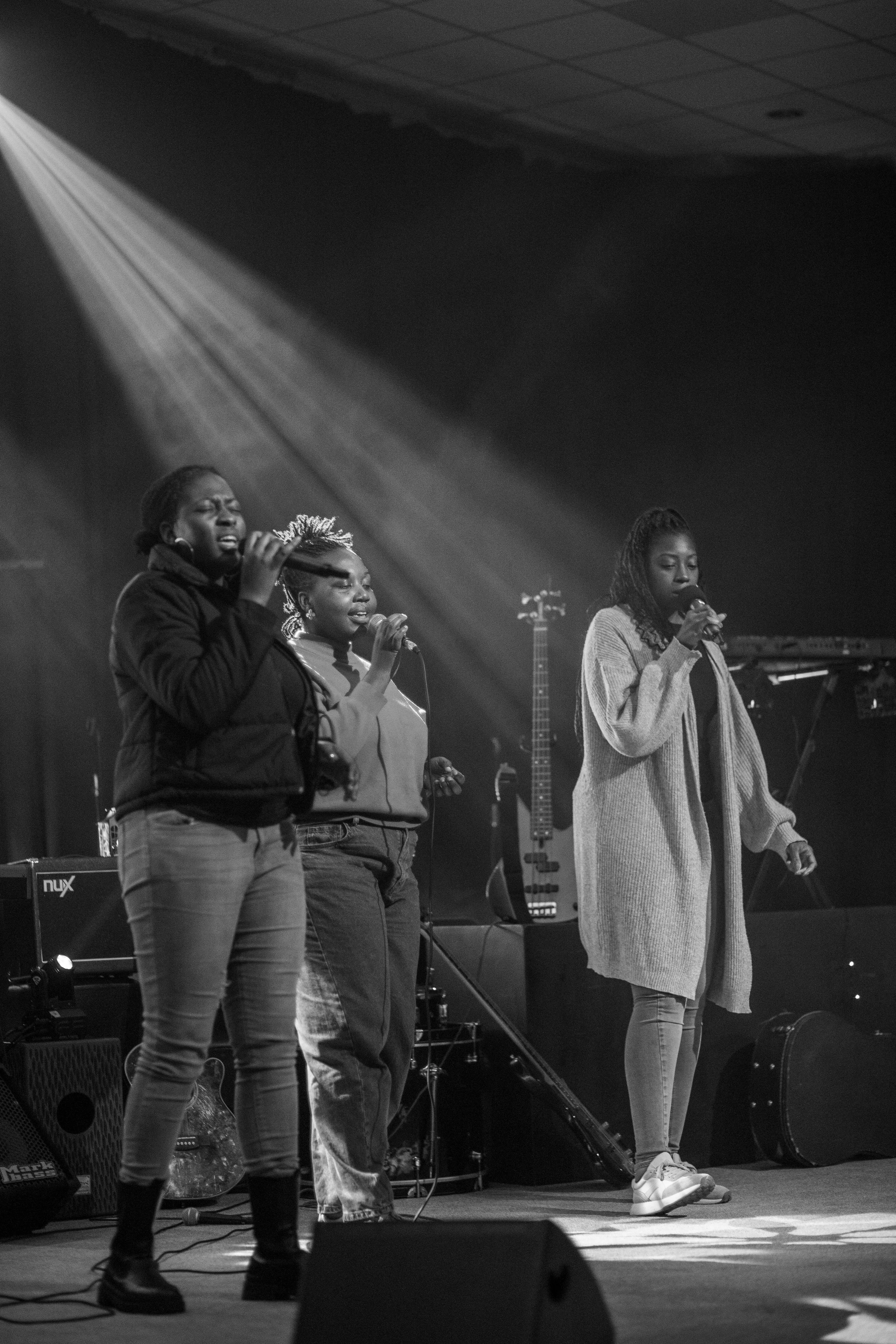 Three women singing on stage with musical instruments and equipment behind them, in a performance setting.
