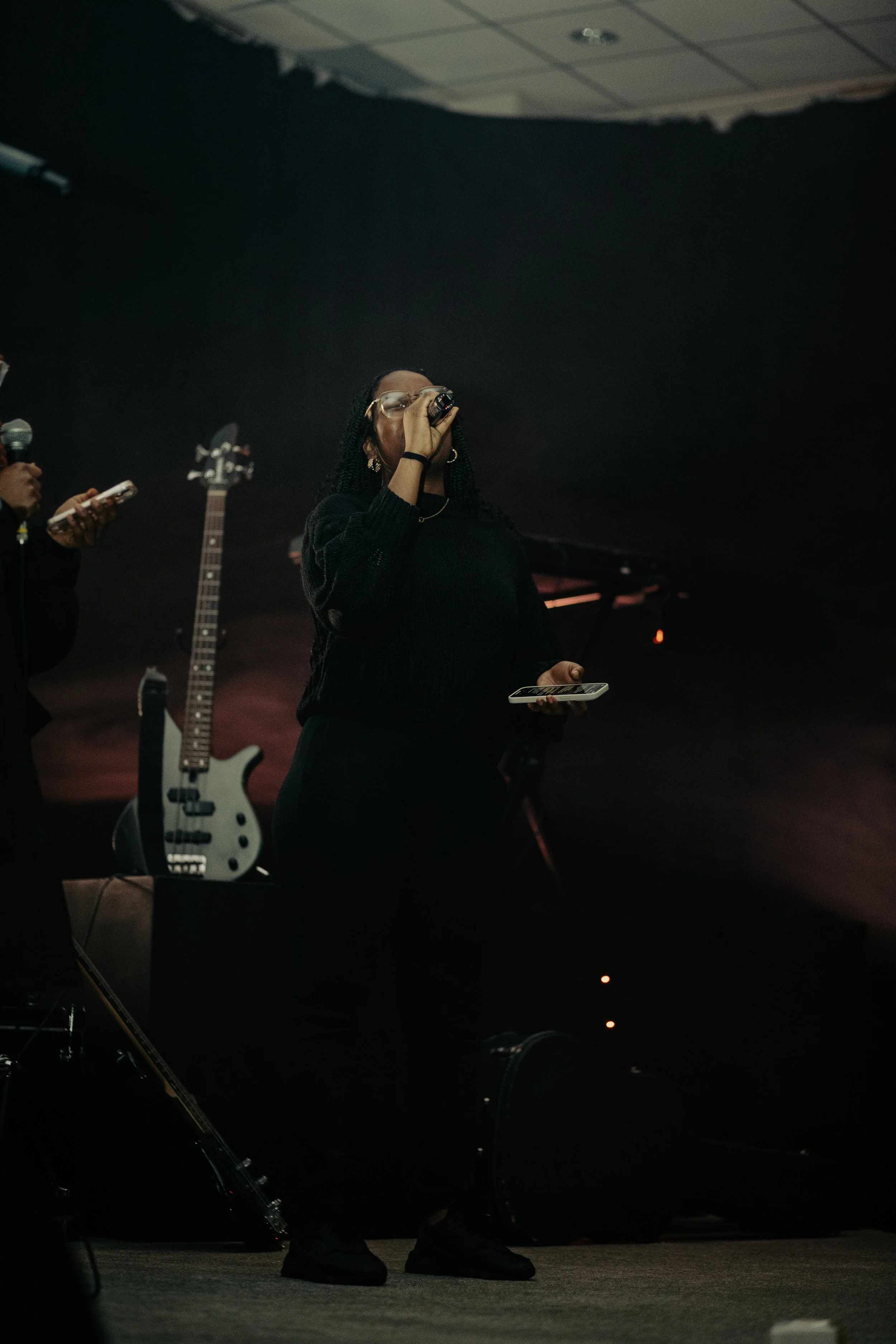 A woman with glasses, wearing a dark sweater, holding a microphone and a smartphone, standing on stage with a black background, a guitar on a stand behind her, and a person on her left checking a device.