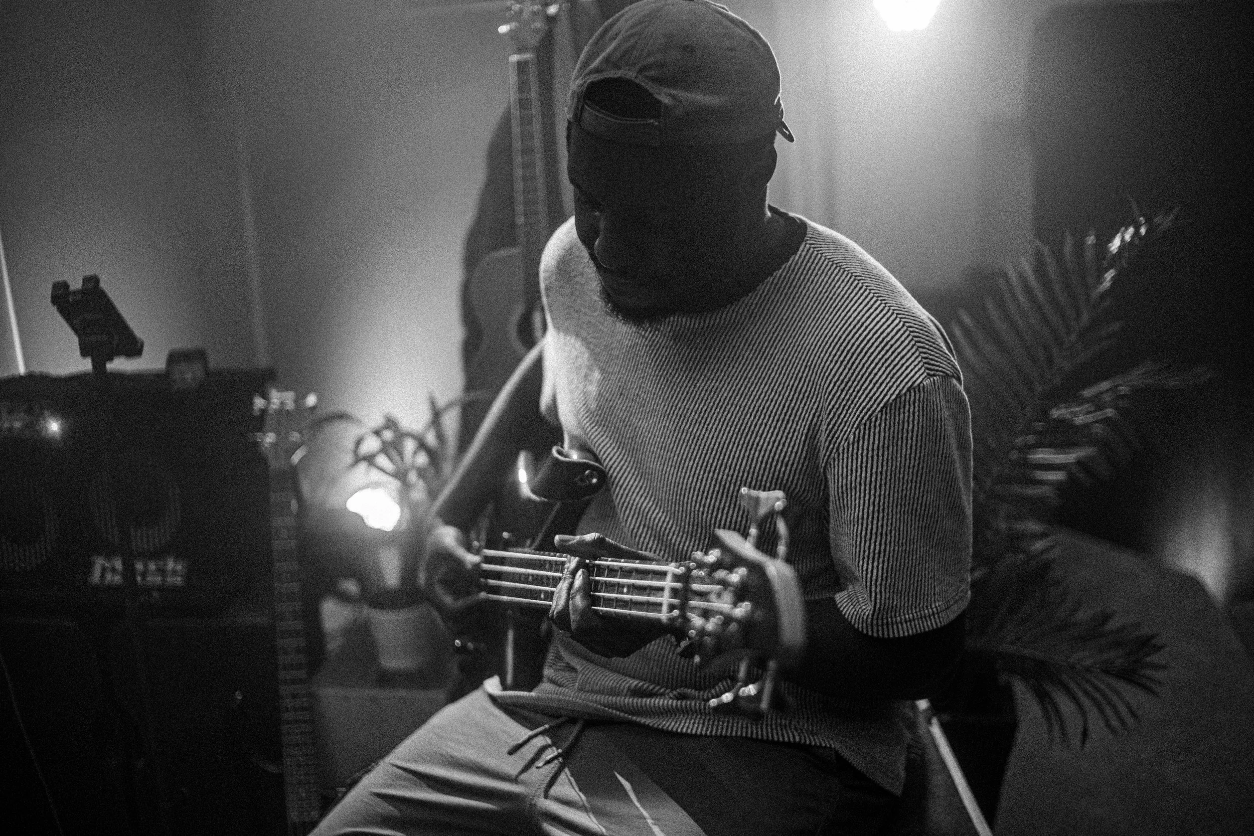 A person wearing a cap and a striped shirt, sitting and playing an acoustic guitar in a dimly lit room decorated with guitars and plants.