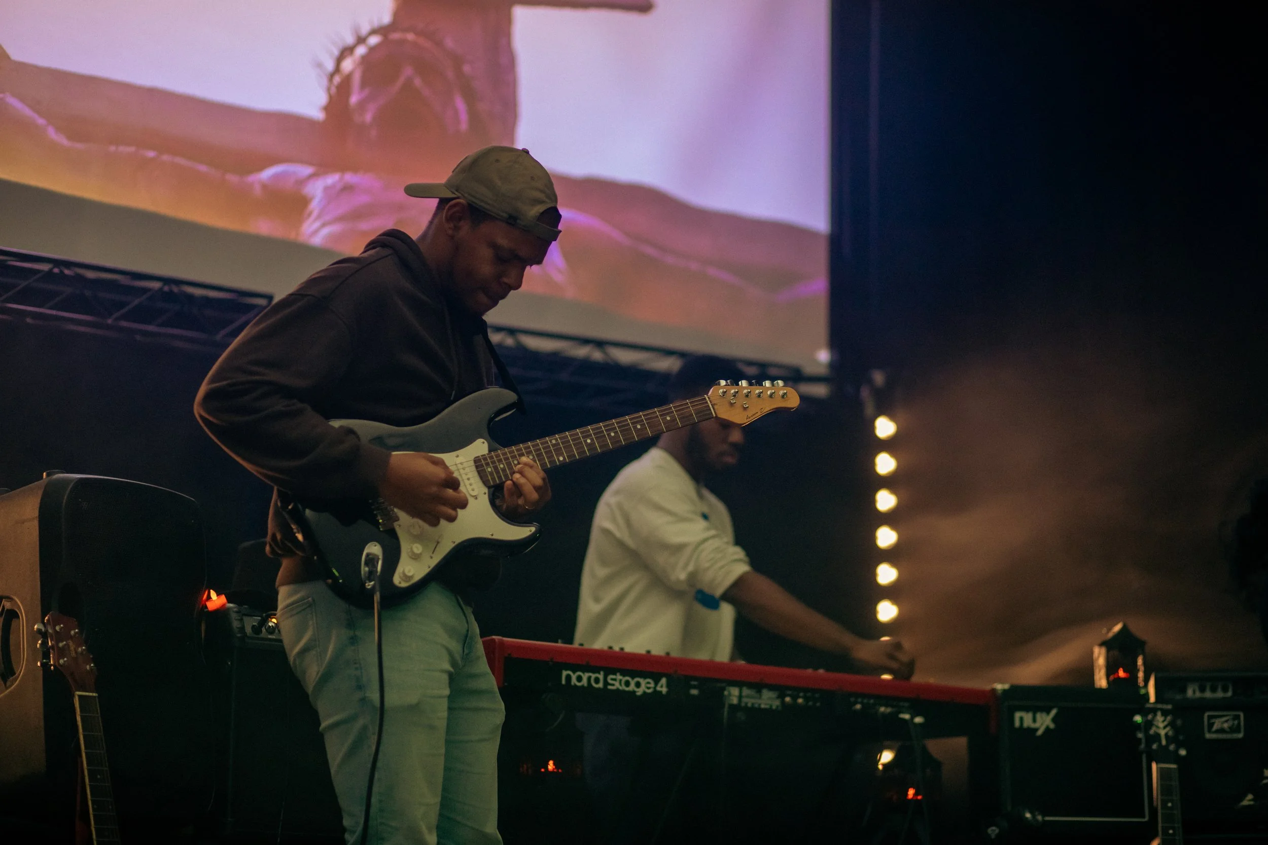 Musicians performing on stage, including a guitarist in a backwards cap and another playing a keyboard, with a large screen displaying a crossed-arms person in the background.