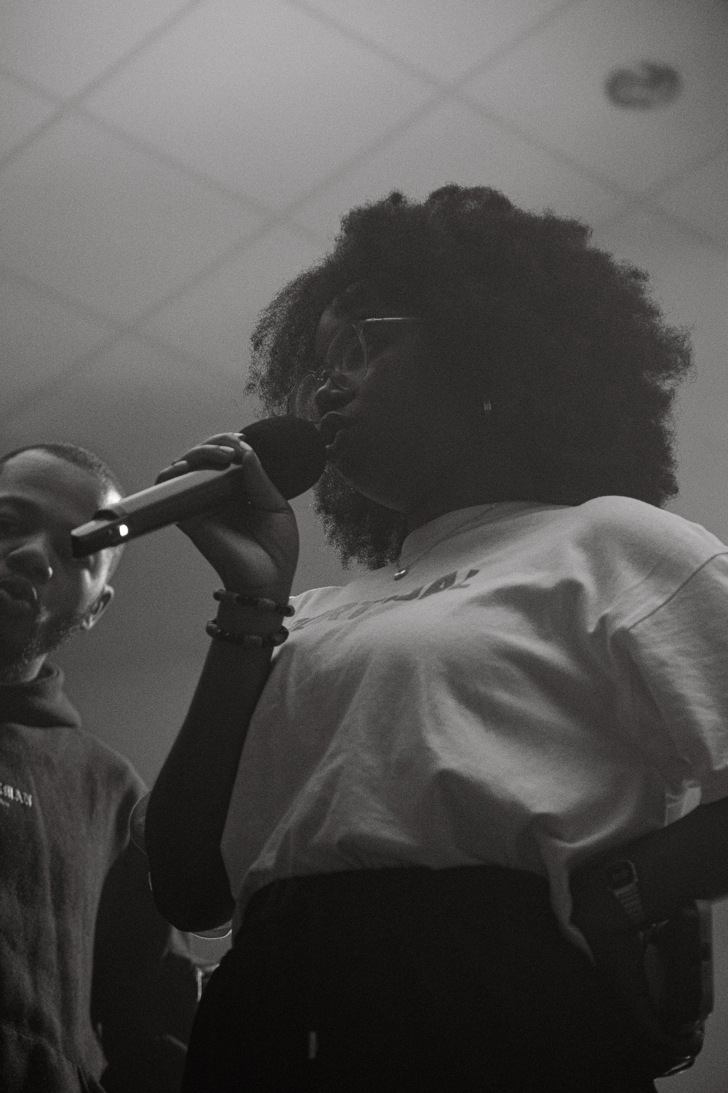 A woman with glasses singing into a microphone, wearing a light-colored shirt and a wristwatch.