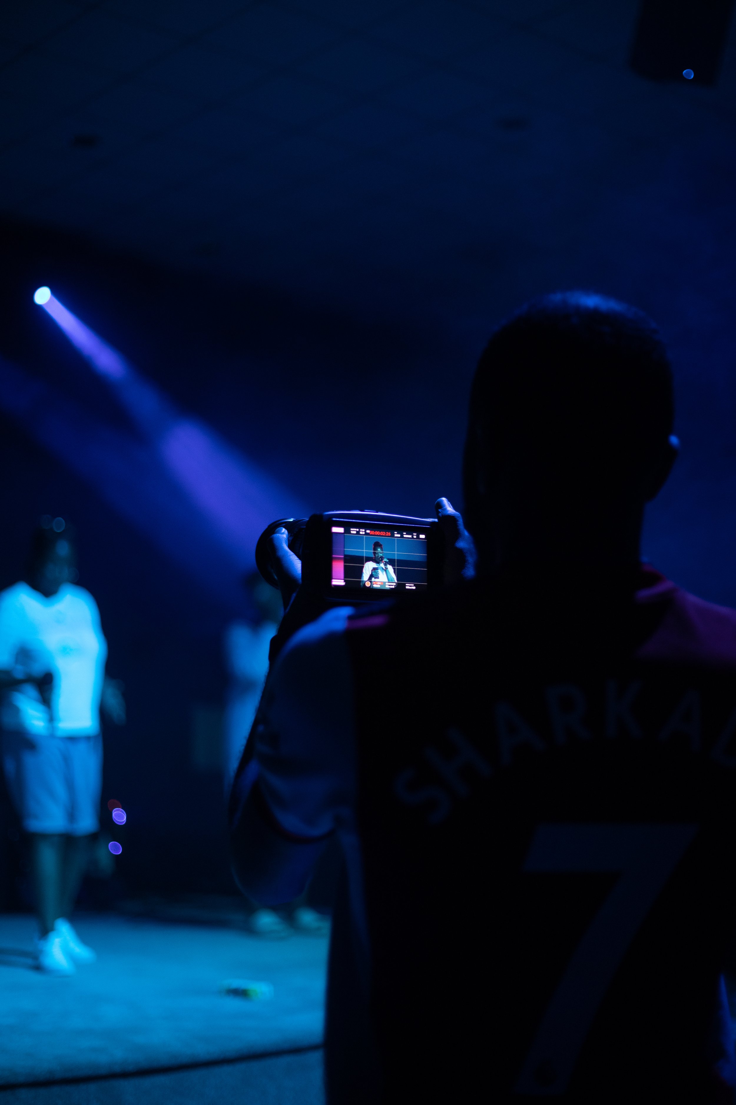 A person holding a camera is taking a photo of a woman standing in a dark room with blue lighting, while another person stands in the background.
