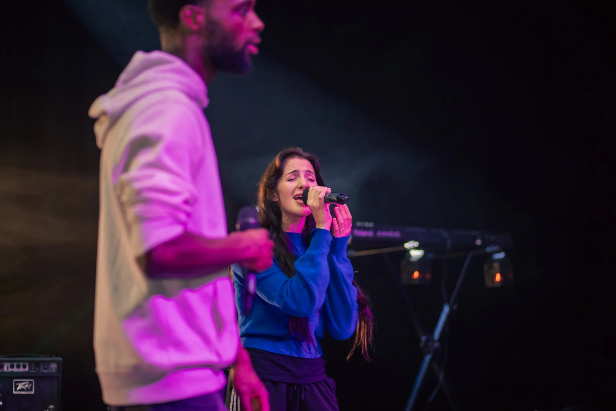 A man in a white hoodie and a woman in a blue top are on stage. The woman is singing into a microphone with her eyes closed. The background is dark with some musical equipment visible.