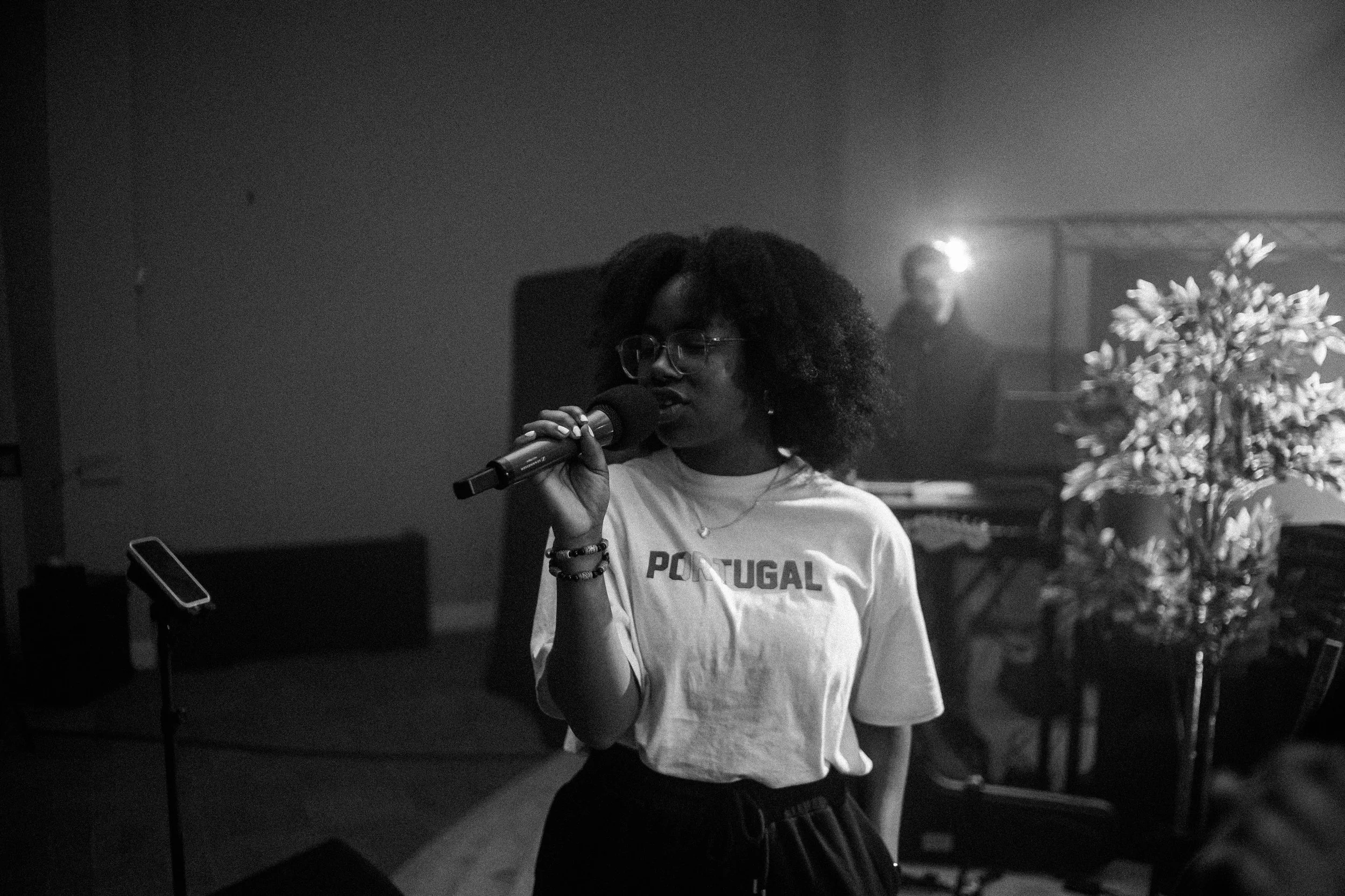 A woman with glasses and curly hair holding a microphone in a room with a plant and a person in the background.