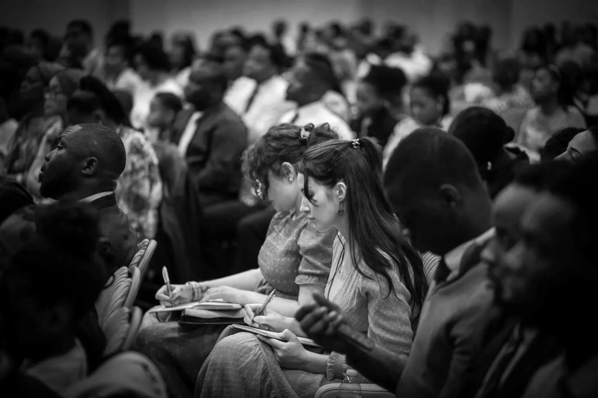 Photo of a diverse group of people attending a conference or seminar, sitting in a large room, some taking notes, with a focus on a woman in traditional attire.