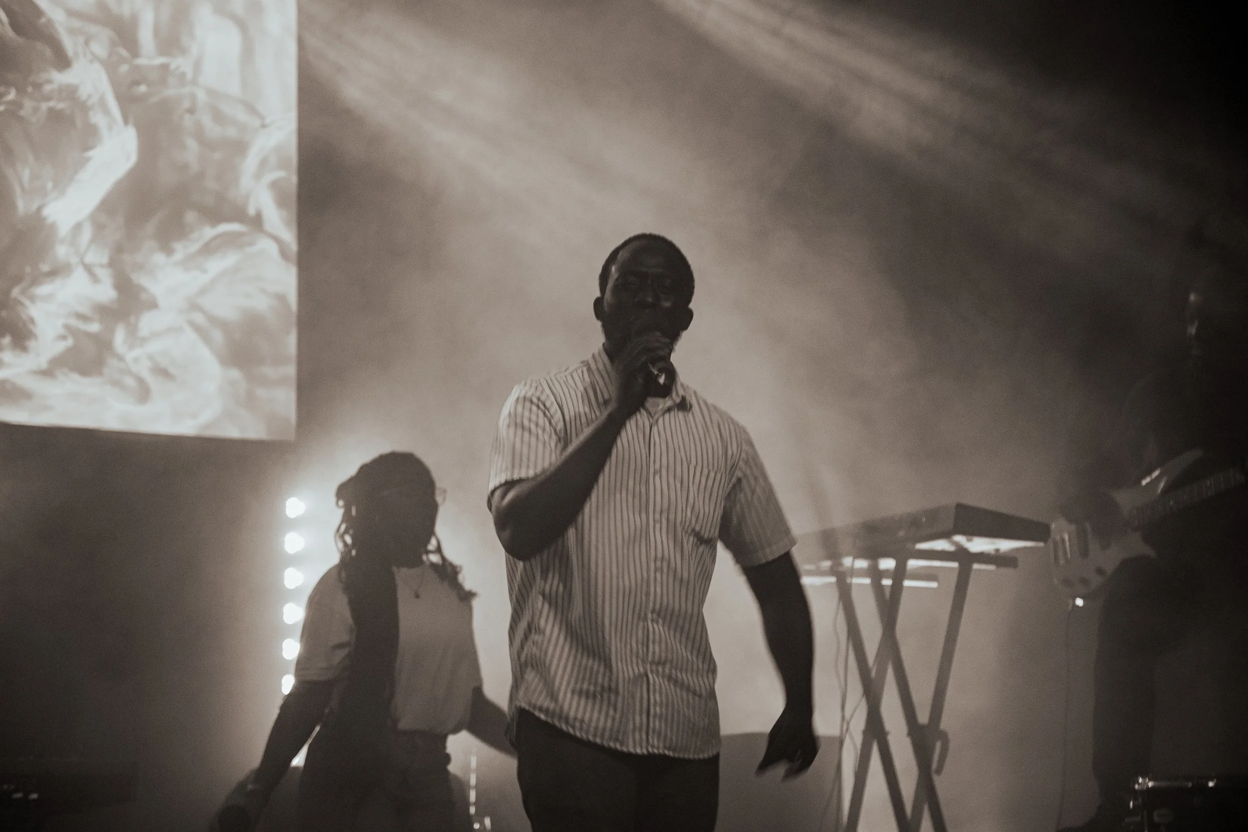 Man in striped shirt holding a microphone, standing in a smoky or foggy room with silhouettes of other people and musical equipment visible in the background.