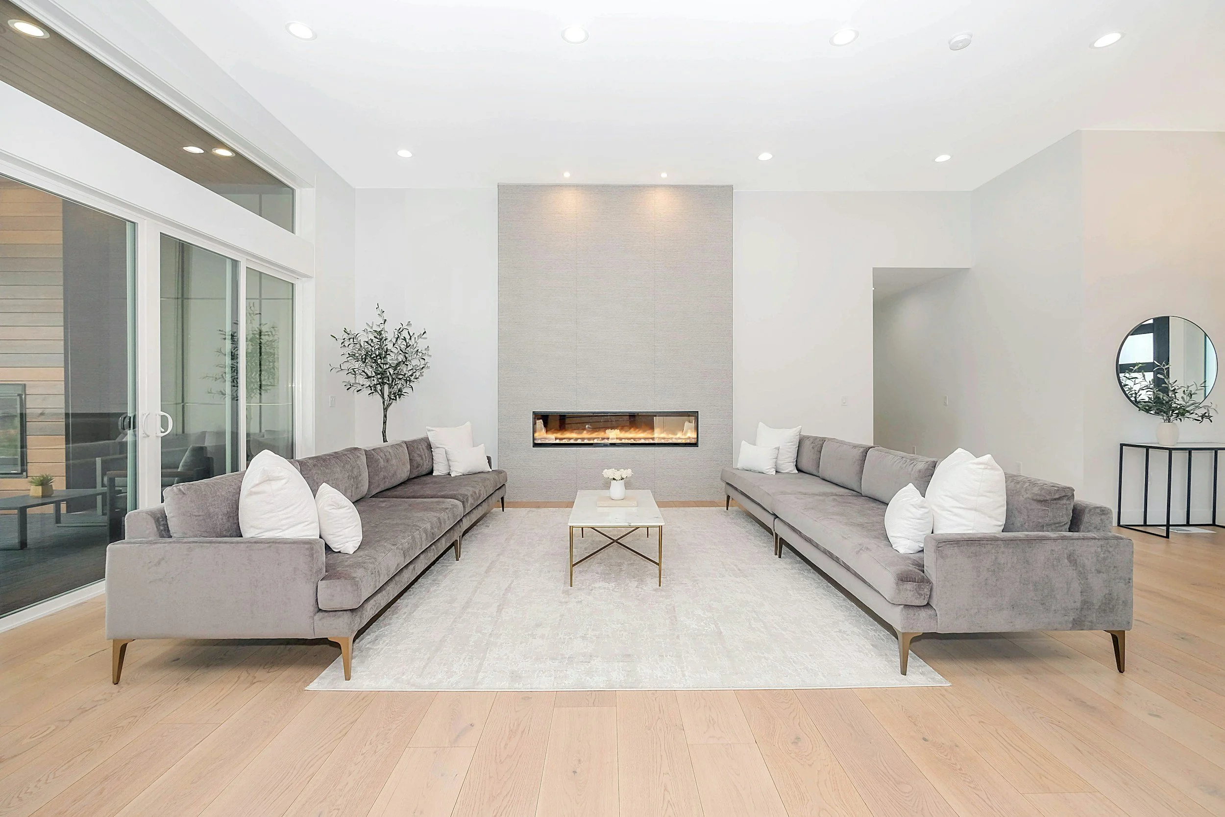 Modern living room with two long gray sofas, white pillows, a white rug, glass sliding door, fireplace, small white table with flowers, and minimal decor.