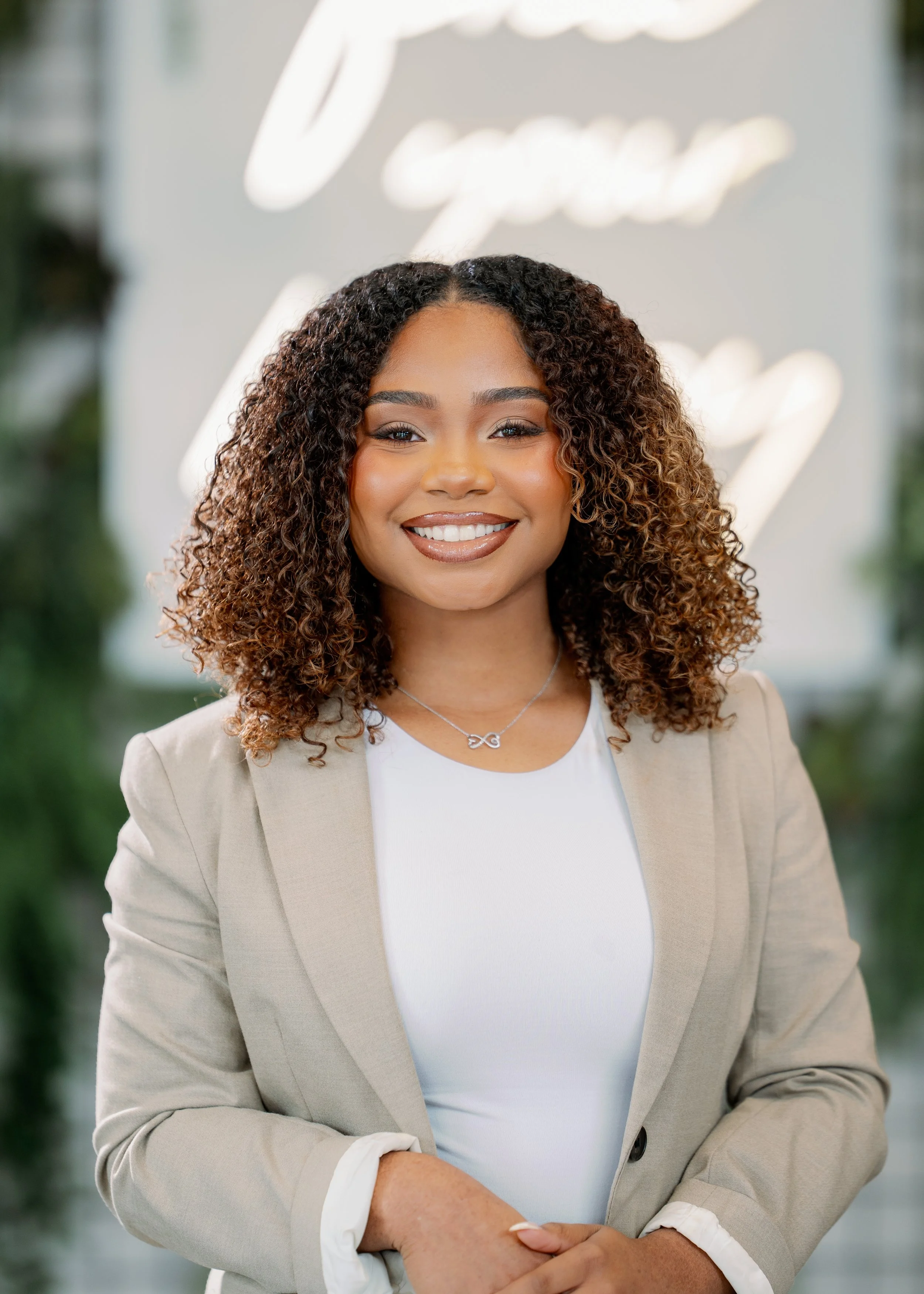 A smiling woman with curly hair, wearing a beige blazer over a white top, standing in front of a blurred background with green plants and a neon sign.