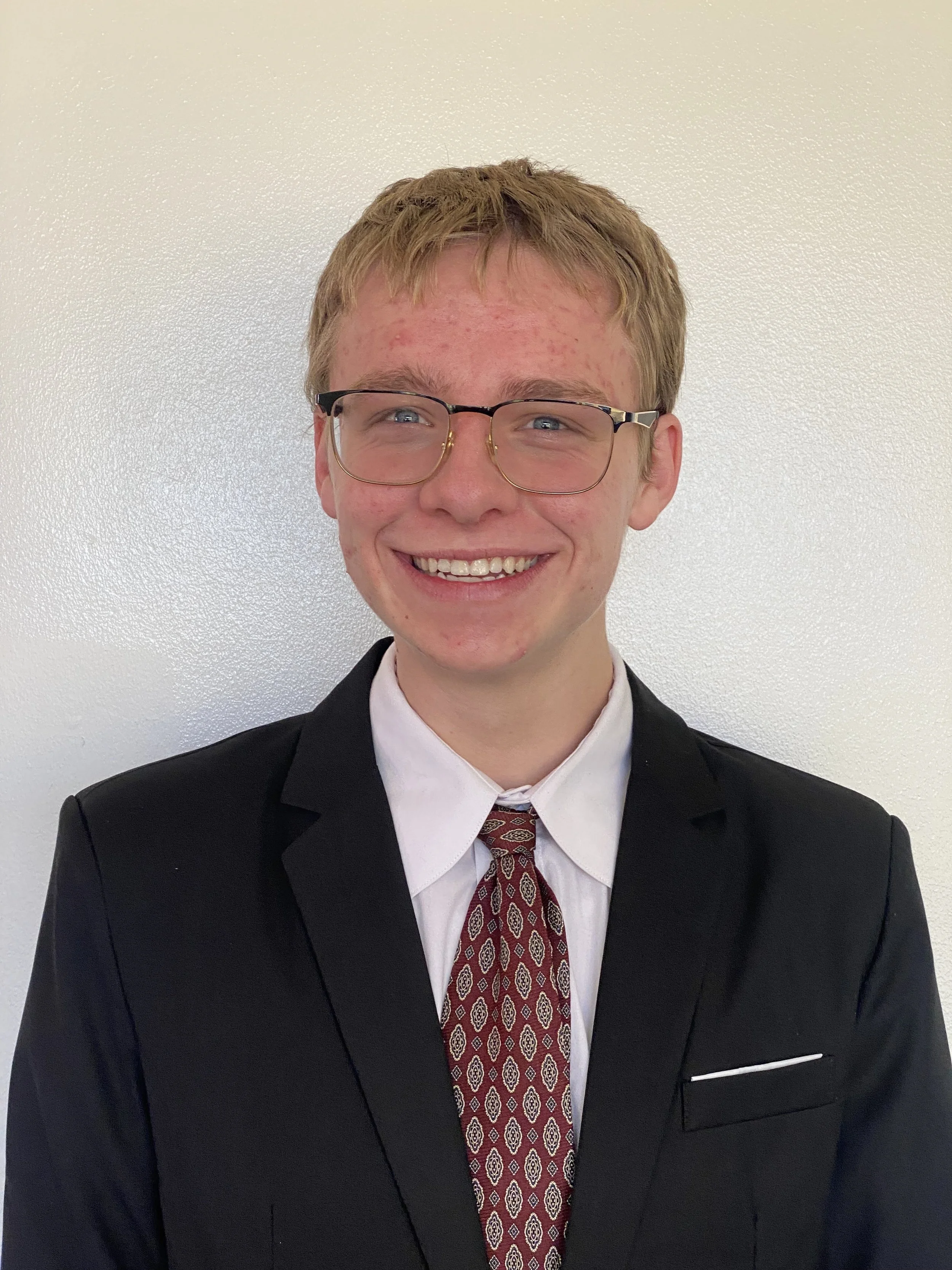 Young man with glasses, short blond hair, and red acne, wearing a black suit, white shirt, and patterned red tie, smiling against a plain white wall.