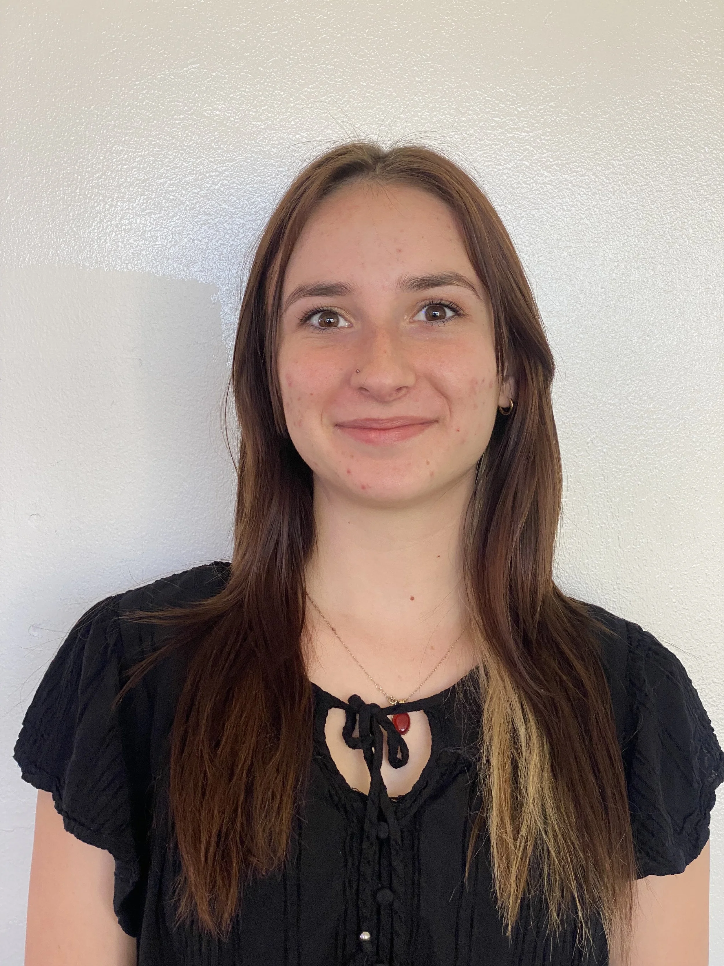 A young woman with brown hair, wearing a black top and a necklace, standing in front of a plain white wall, smiling.