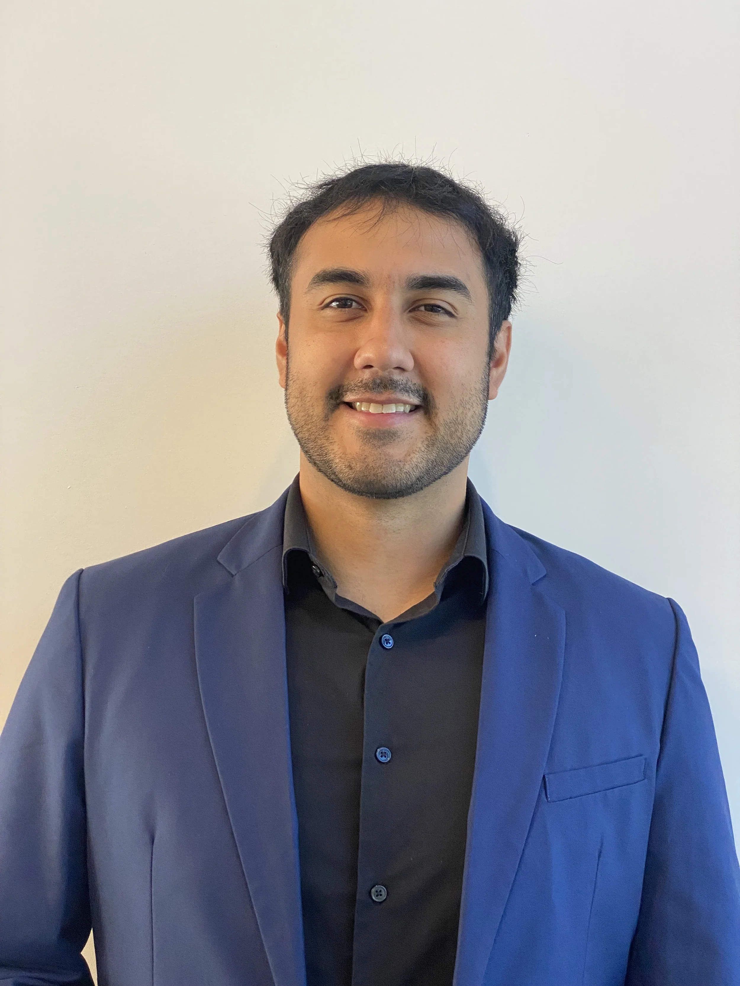 A man with dark hair and a beard wearing a black shirt and a blue blazer, standing against a plain light-colored background, smiling at the camera.