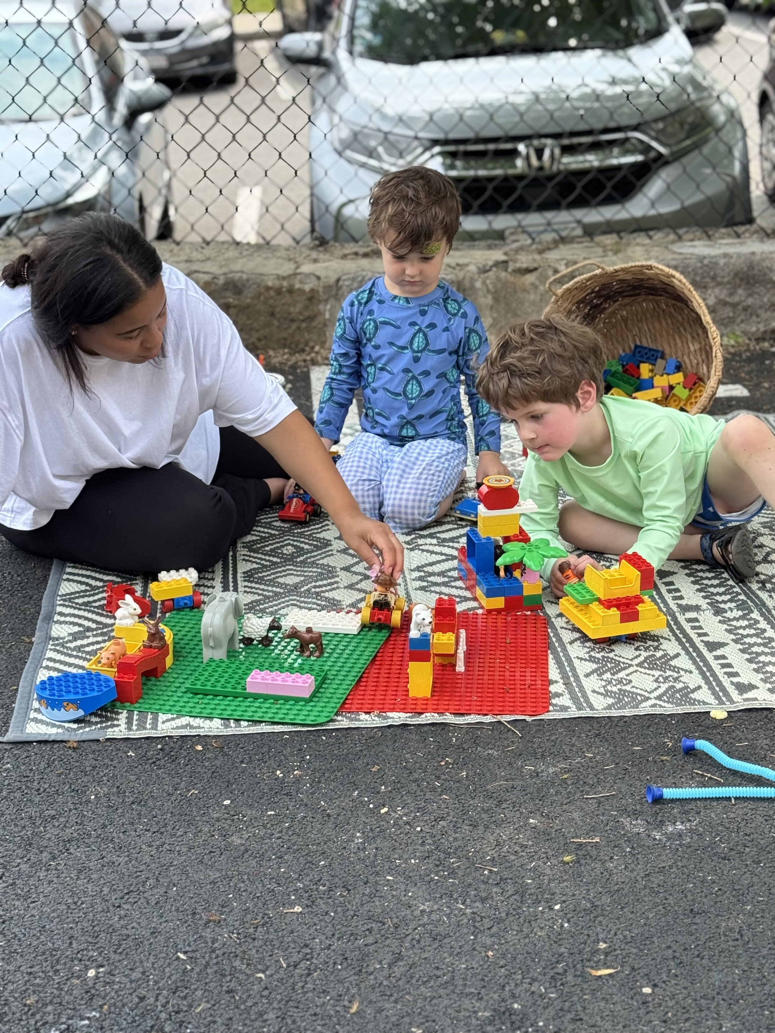 Three children and an adult sitting on a patterned rug outdoors, playing with colorful LEGO bricks. There are cars and a chain-link fence in the background.