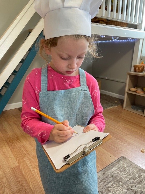 A young girl wearing a chef's hat, a pink shirt with heart patterns, and a blue apron, standing and writing on a clipboard with a pencil in a cozy indoor setting.