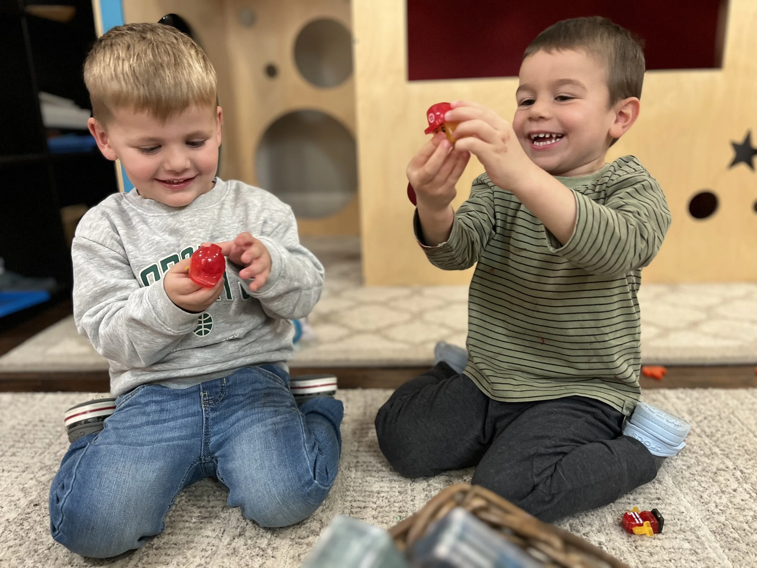 Two young boys sitting on a carpeted floor playing with small plastic toys, smiling and laughing, with a wooden play structure in the background.