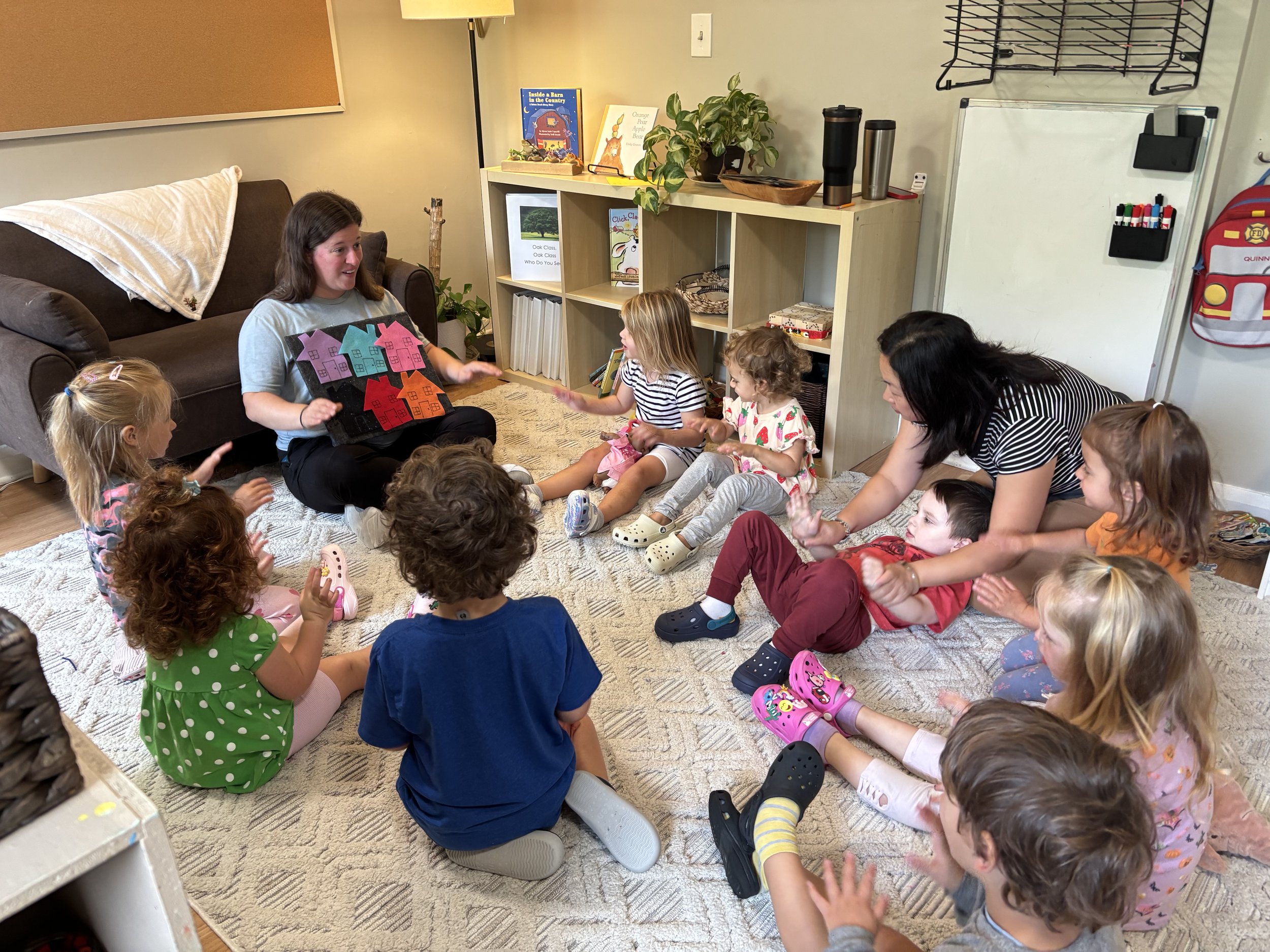 Children and two adults sitting on a carpet in a circle, engaging in a storytelling or educational activity in a cozy room.
