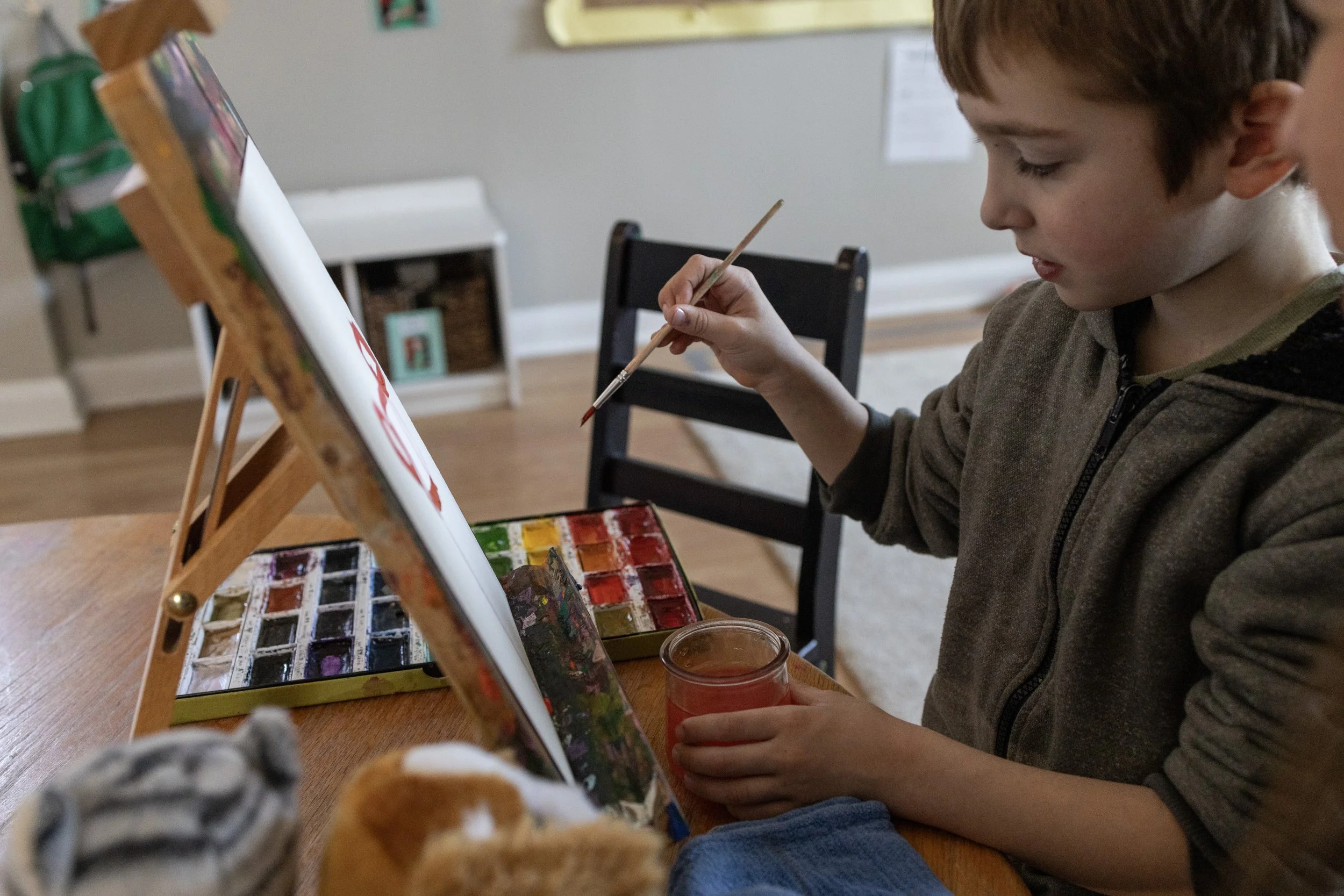 A young boy is painting with watercolor paints at a table, holding a brush and a glass of water, with more paint supplies nearby.