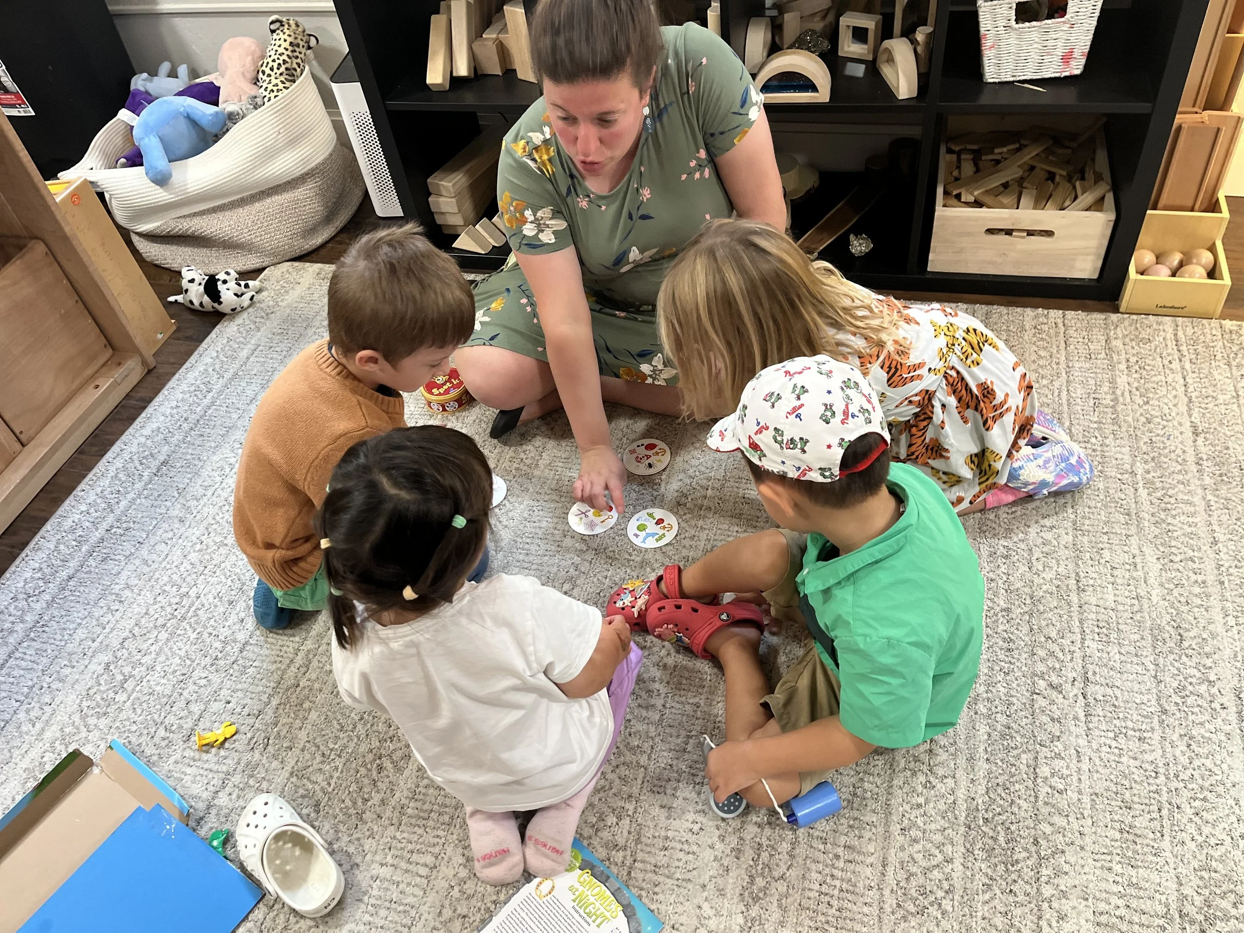 A woman and four children sitting on a carpeted floor, playing a tabletop game with circular cards. The woman and girl are in cartoon-character pajamas, and the children are dressed in casual clothes. Toys and a storage shelf with wooden blocks and baskets are visible in the background.