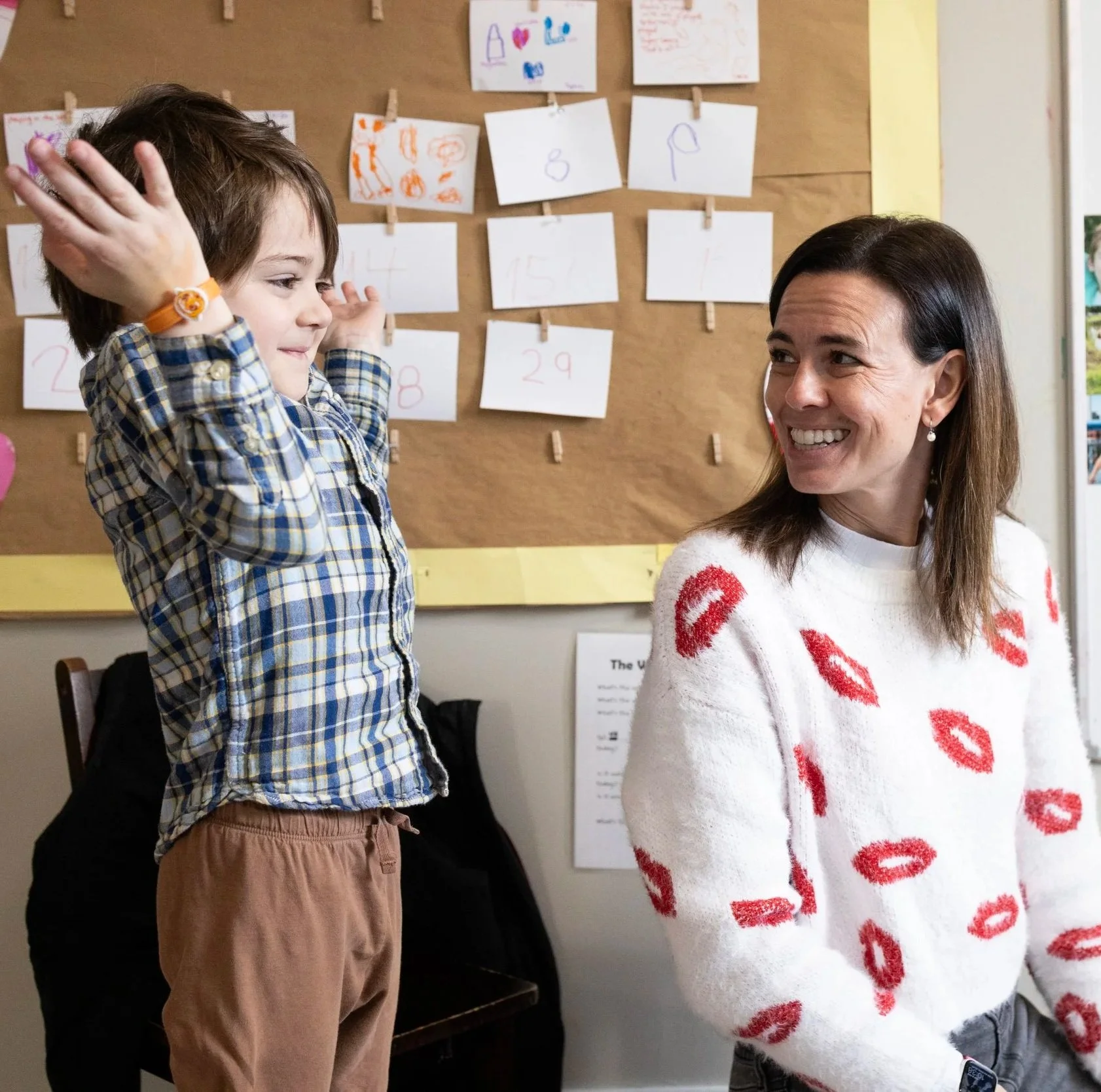 A young boy and a woman are in a classroom. The boy appears excited with his arms raised, and the woman is smiling at him. Behind them is a bulletin board with children's drawings and numbers.