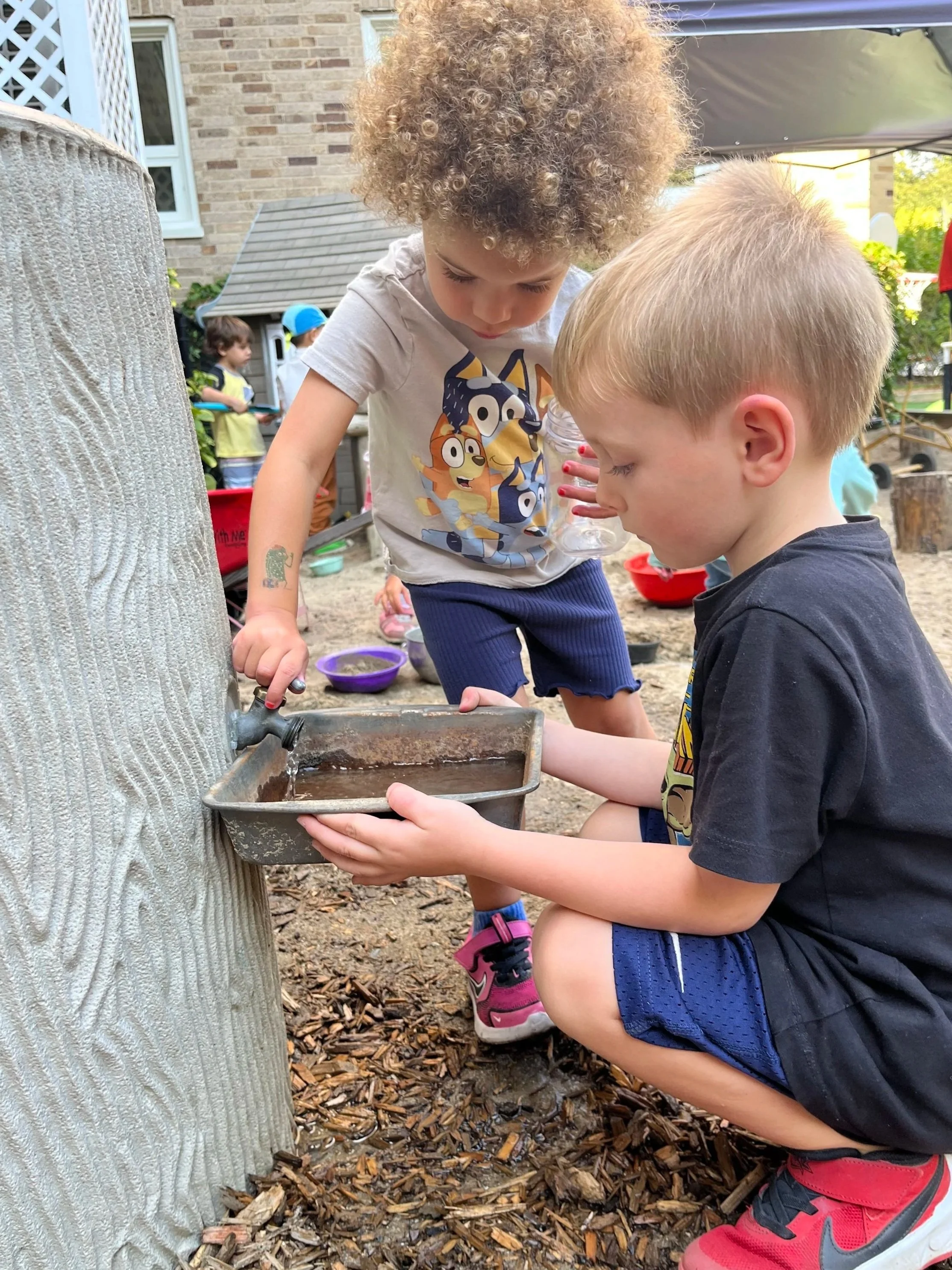 Two young children, a girl with curly hair and a boy with short blonde hair, are playing outdoors with a water fountain attached to a tree. The girl is pouring water into a tray held by the boy. Other children can be seen in the background in a playground area.