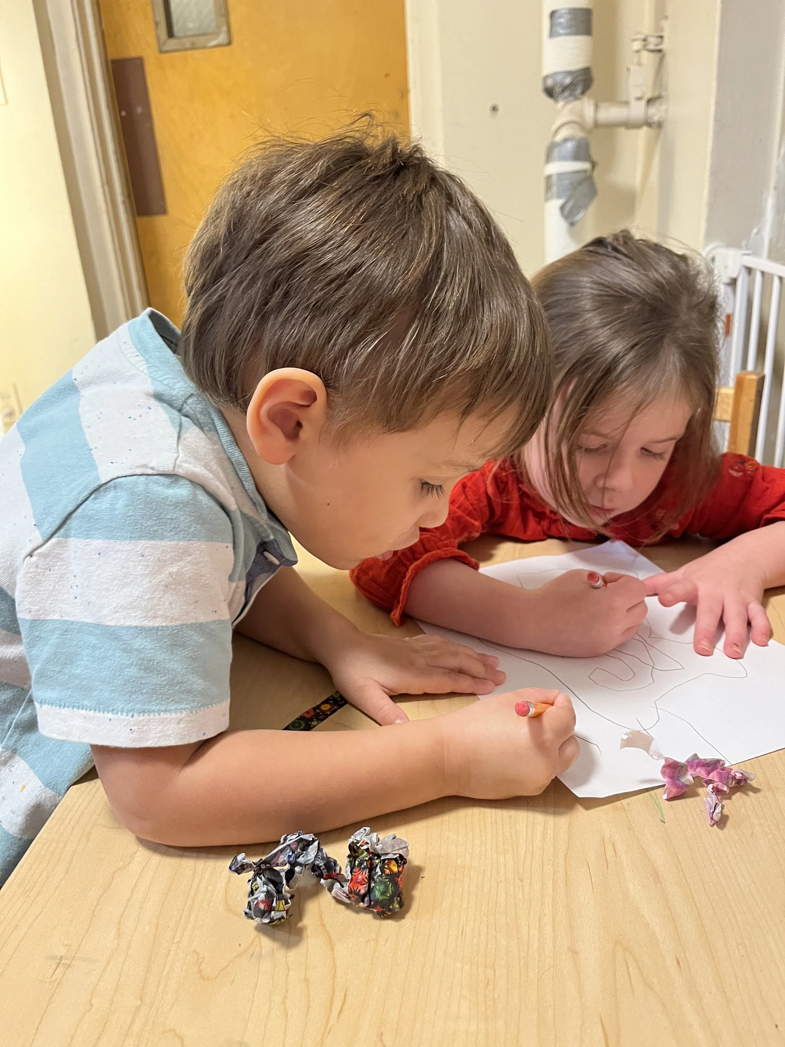 Two children are drawing on a piece of paper at a table, with crumpled pieces of paper nearby.