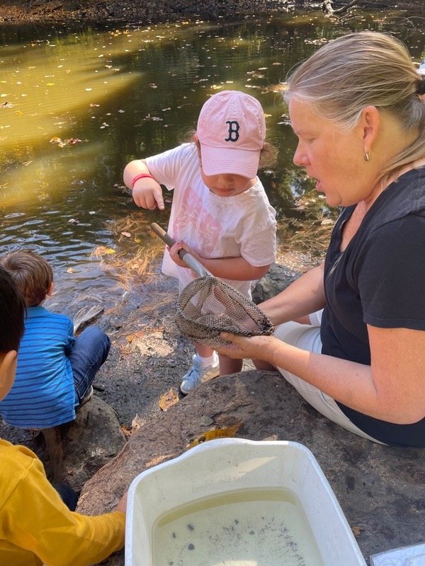 A woman and a young girl look into a small mesh net held over a river, possibly for catching fish or exploring aquatic life. Other children sit nearby on rocks.