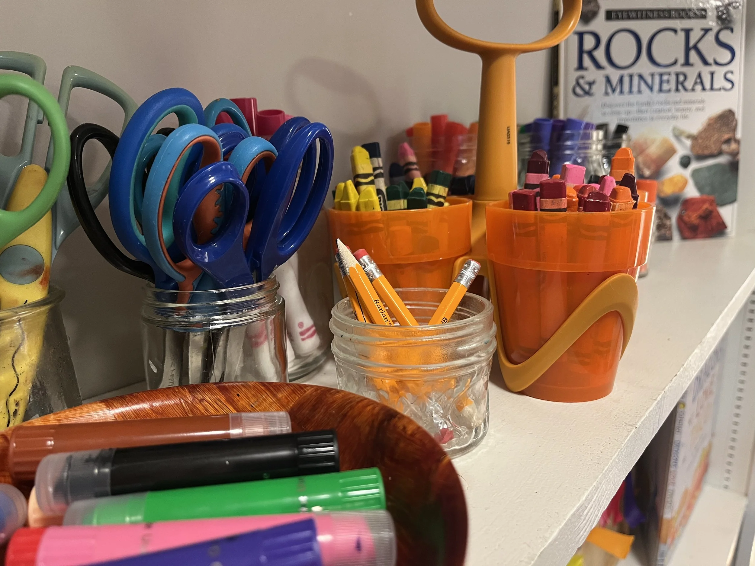 A shelf with various stationery supplies including colorful scissors, pencils, and markers, along with a book titled『Rocks & Minerals』 in the background.