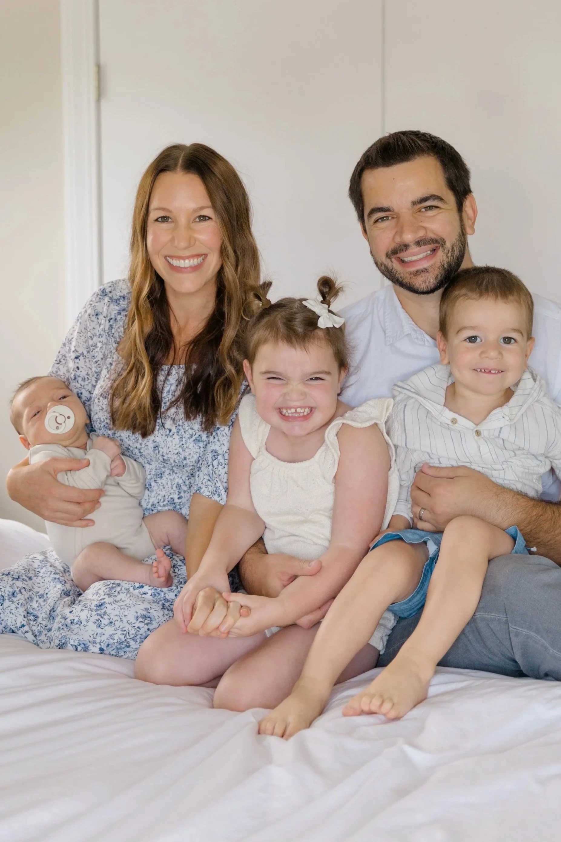 A smiling family of five sitting on a bed, with two parents and three children, two girls and a boy, in a well-lit room.
