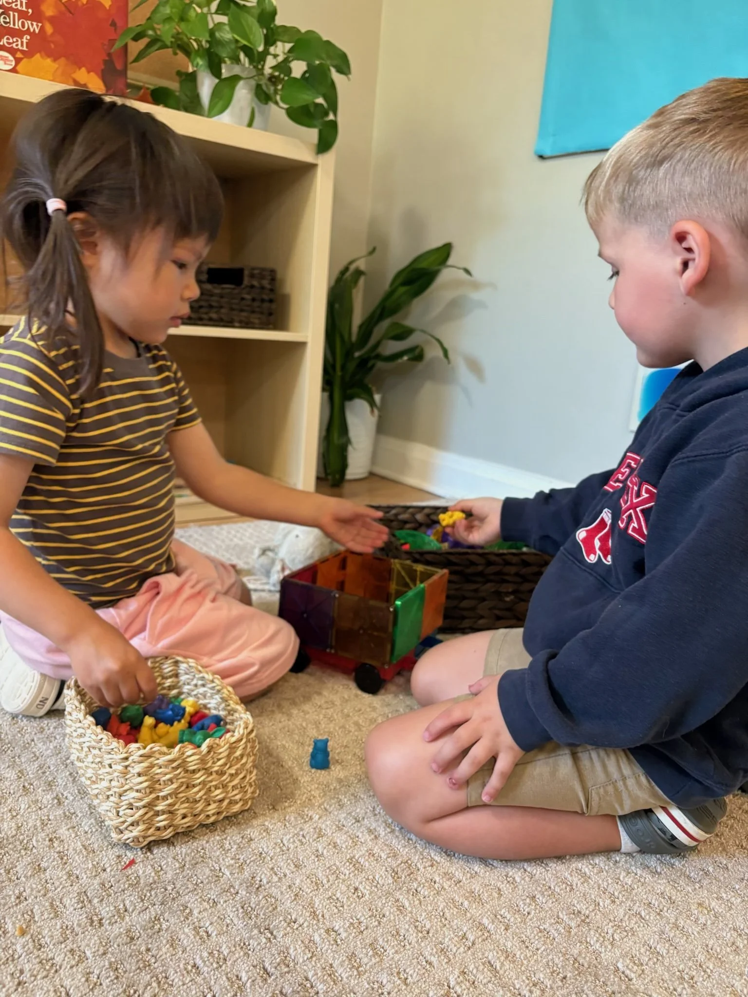 Two children playing with colorful toys on a beige carpet in a room with a bookshelf, plants, and a blue wall hanging.
