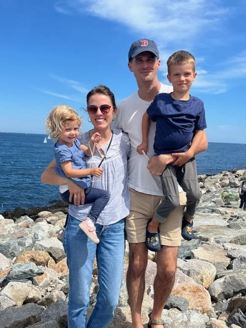A family of four stands on a rocky shoreline with the ocean and a sailboat in the background. The woman is holding a young girl and the man is holding a young boy; all are smiling and dressed casually.