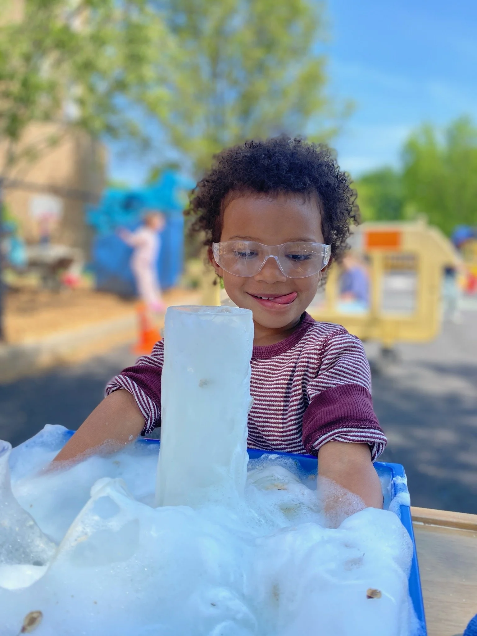 Child wearing safety glasses and a striped shirt playing with ice and bubbles outdoors on a sunny day.