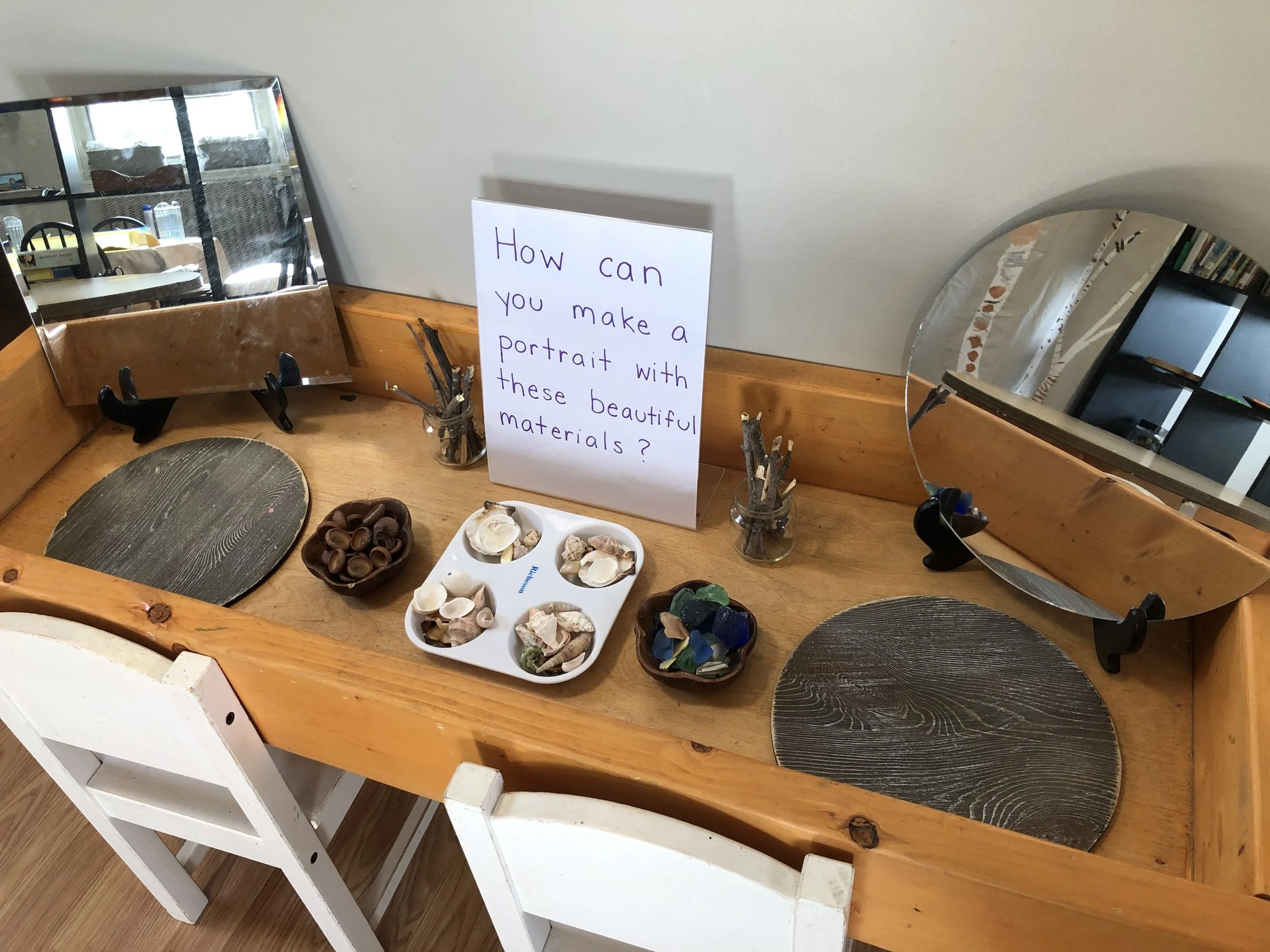 A wooden table with three small mirrors, two marked with black stands, and several shells and rocks for a shell craft activity. A sign with purple handwritten text asks, "How can you make a portrait with these beautiful materials?"