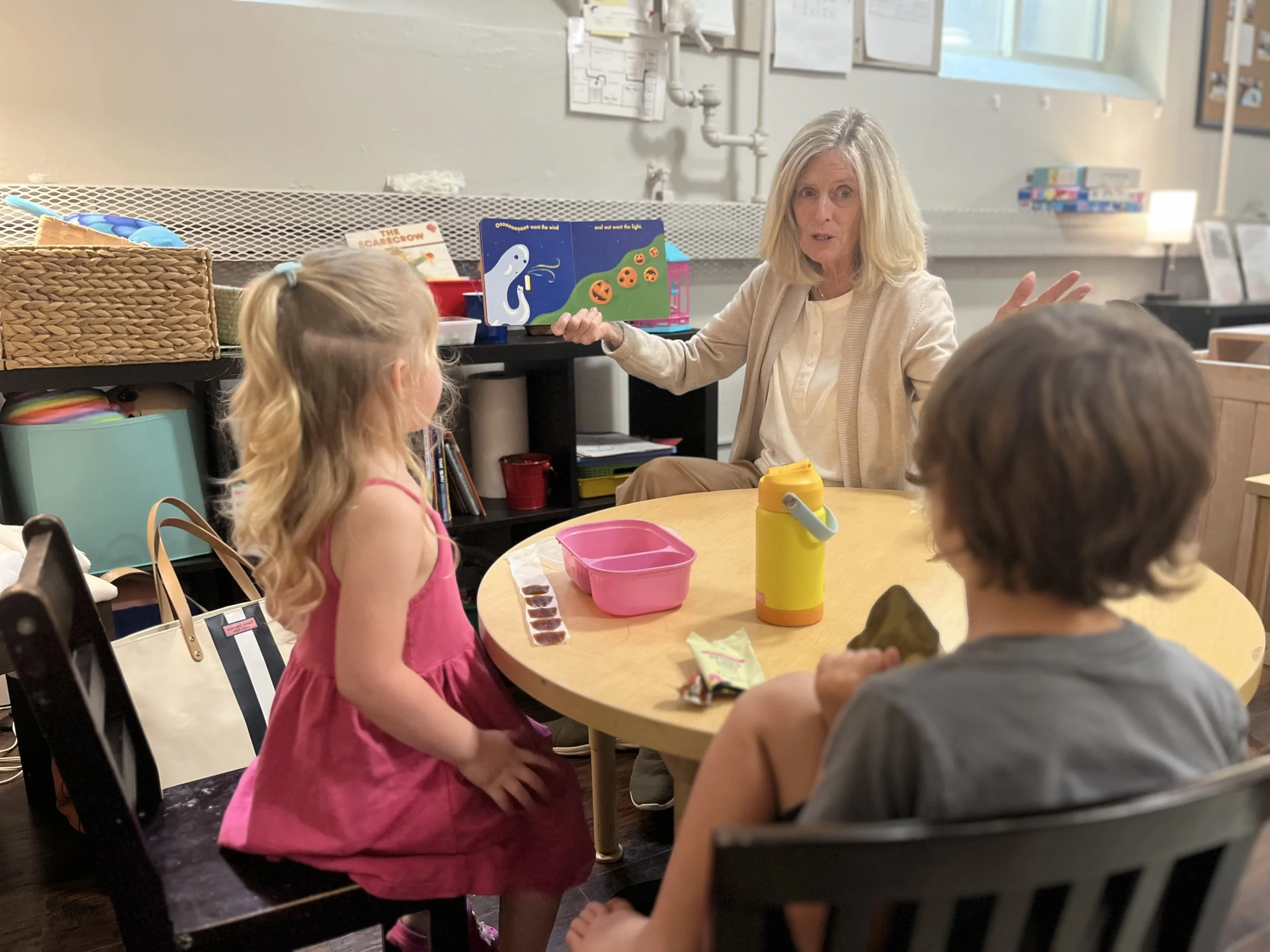 A woman with blonde hair sitting at a table with two children, a girl in a pink dress and a boy with brown hair, in a classroom or daycare setting. The woman appears to be speaking or explaining something, while the children listen. There are toys, books, and a pink lunchbox on the table.