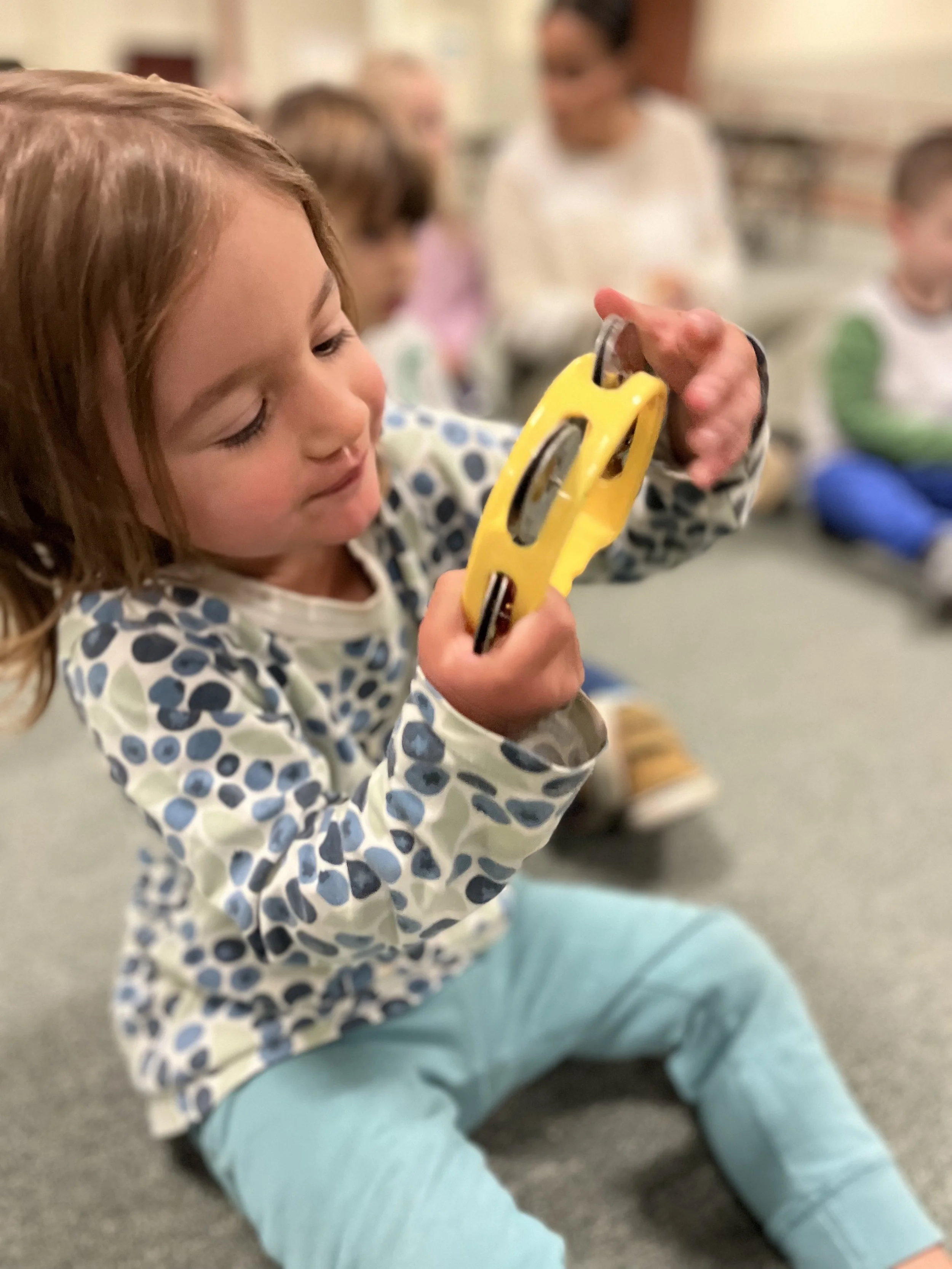 A young girl with brown hair playing with a yellow toy car indoors, sitting on the floor with other children and an adult in the background.