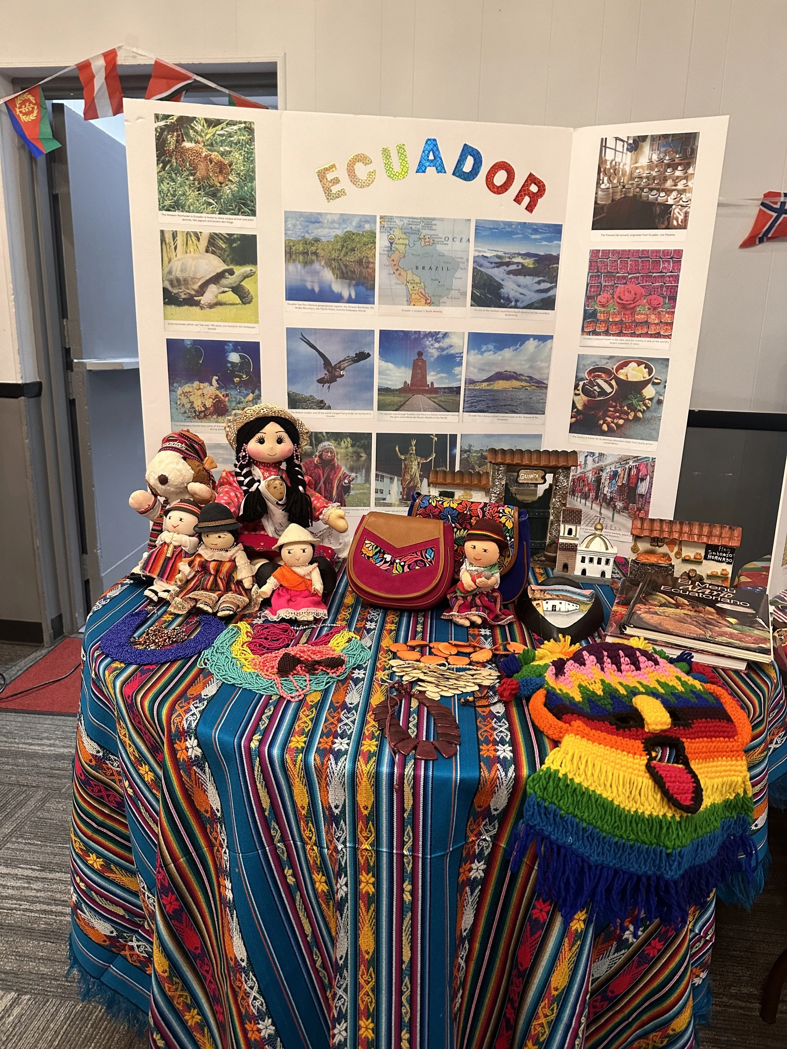 Display of handmade dolls, bags, jewelry, and souvenirs in front of a poster about Ecuador, with photos of landscapes, wildlife, and cultural items of Ecuador.