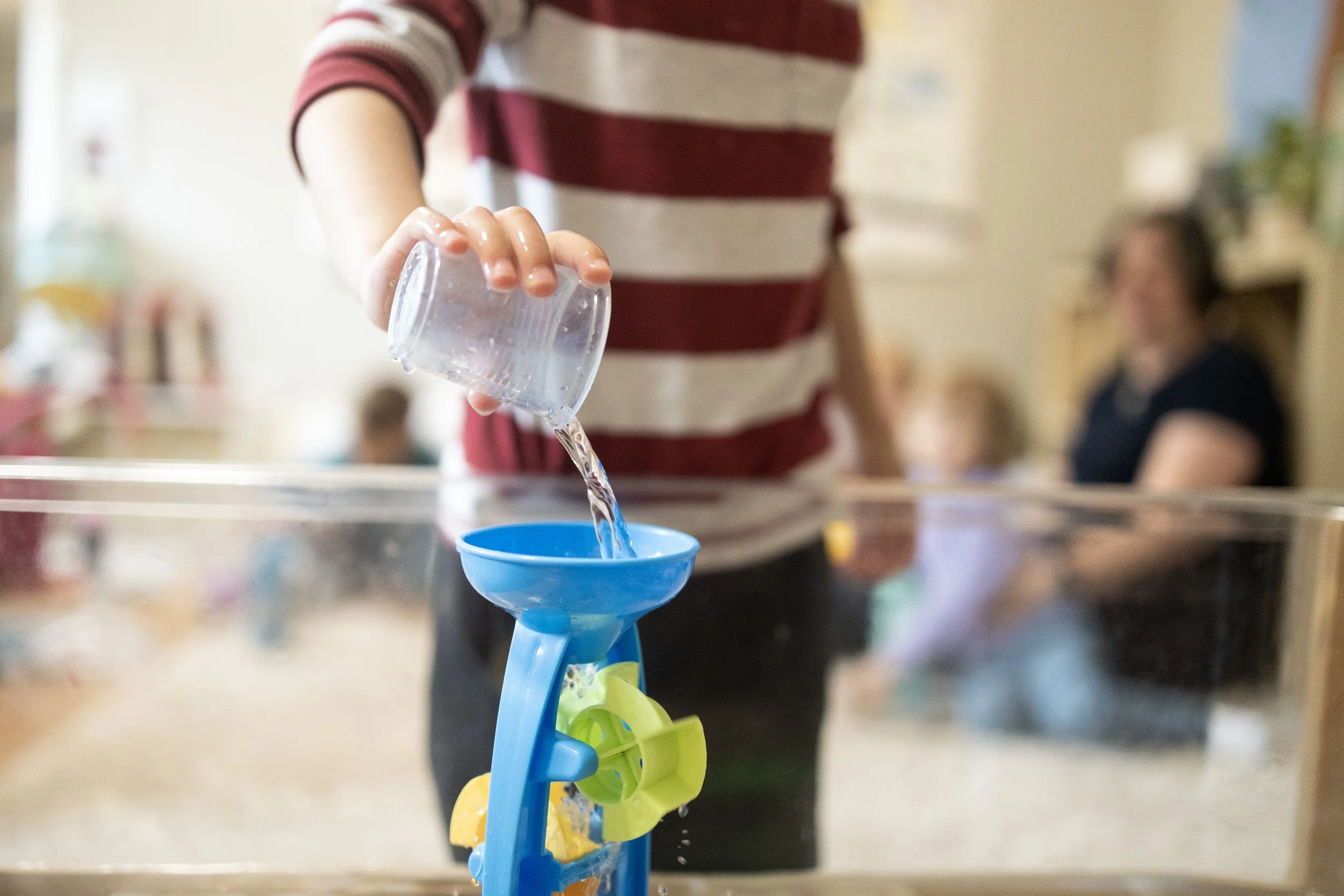 Child pouring water into a blue funnel on a water table, with adults and children blurred in the background.