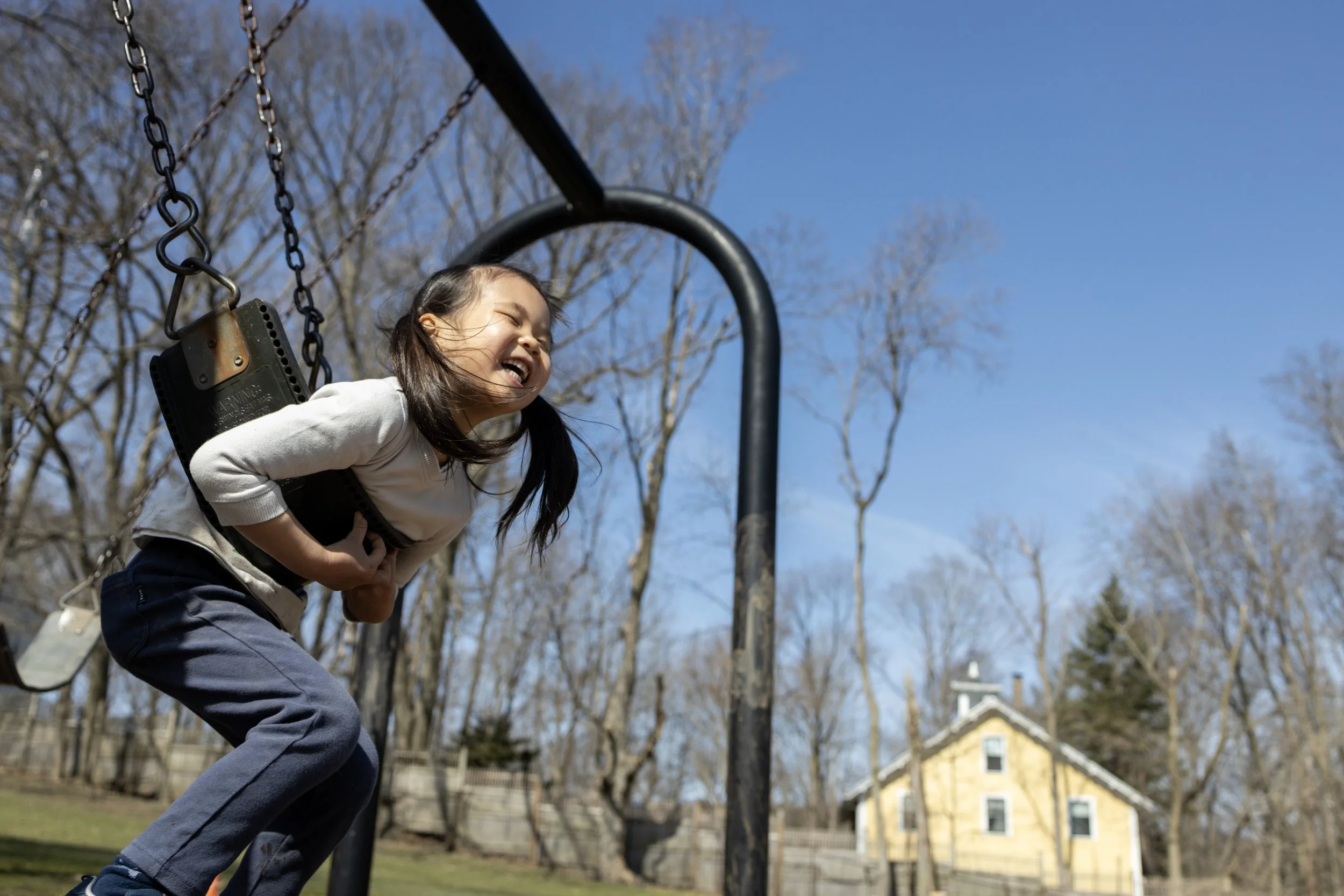 A smiling young girl wearing a white long-sleeve shirt and dark pants playing on a swing set outdoors with a clear blue sky, leafless trees, and a house in the background.
