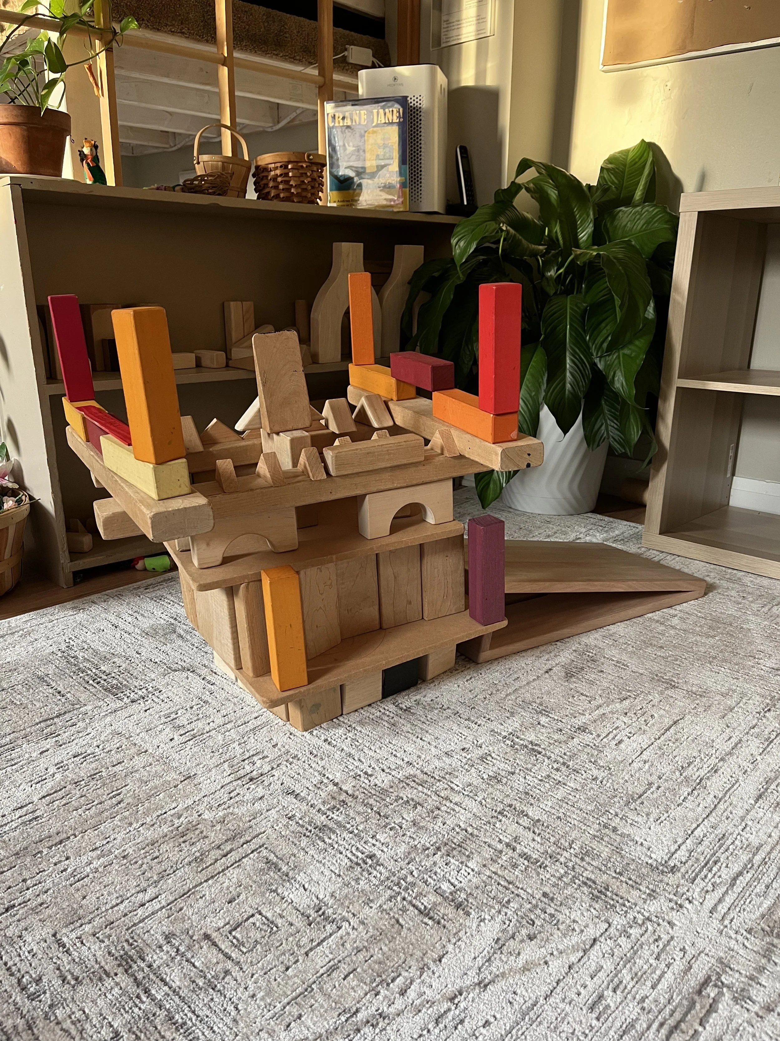 Wooden block stacking game with colorful painted blocks, set up on a wooden floor near a potted plant and in front of a shelf and countertop with baskets and books.