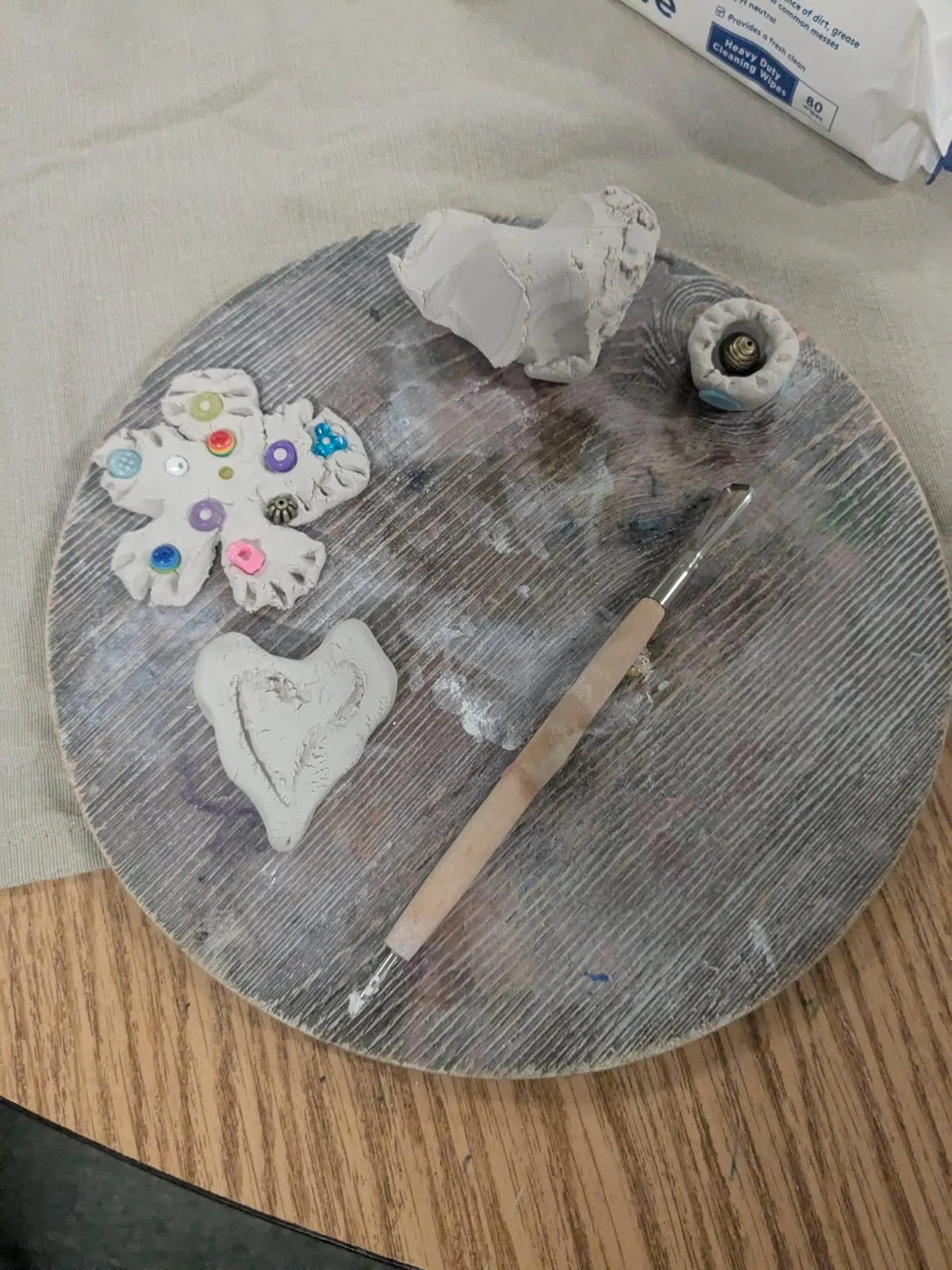 Clay craft project on a wooden table, including clay butterfly with buttons and sequins, a clay heart, and clay bead with a metal component inside. A paintbrush and a bag of clay or plaster are also on the table.