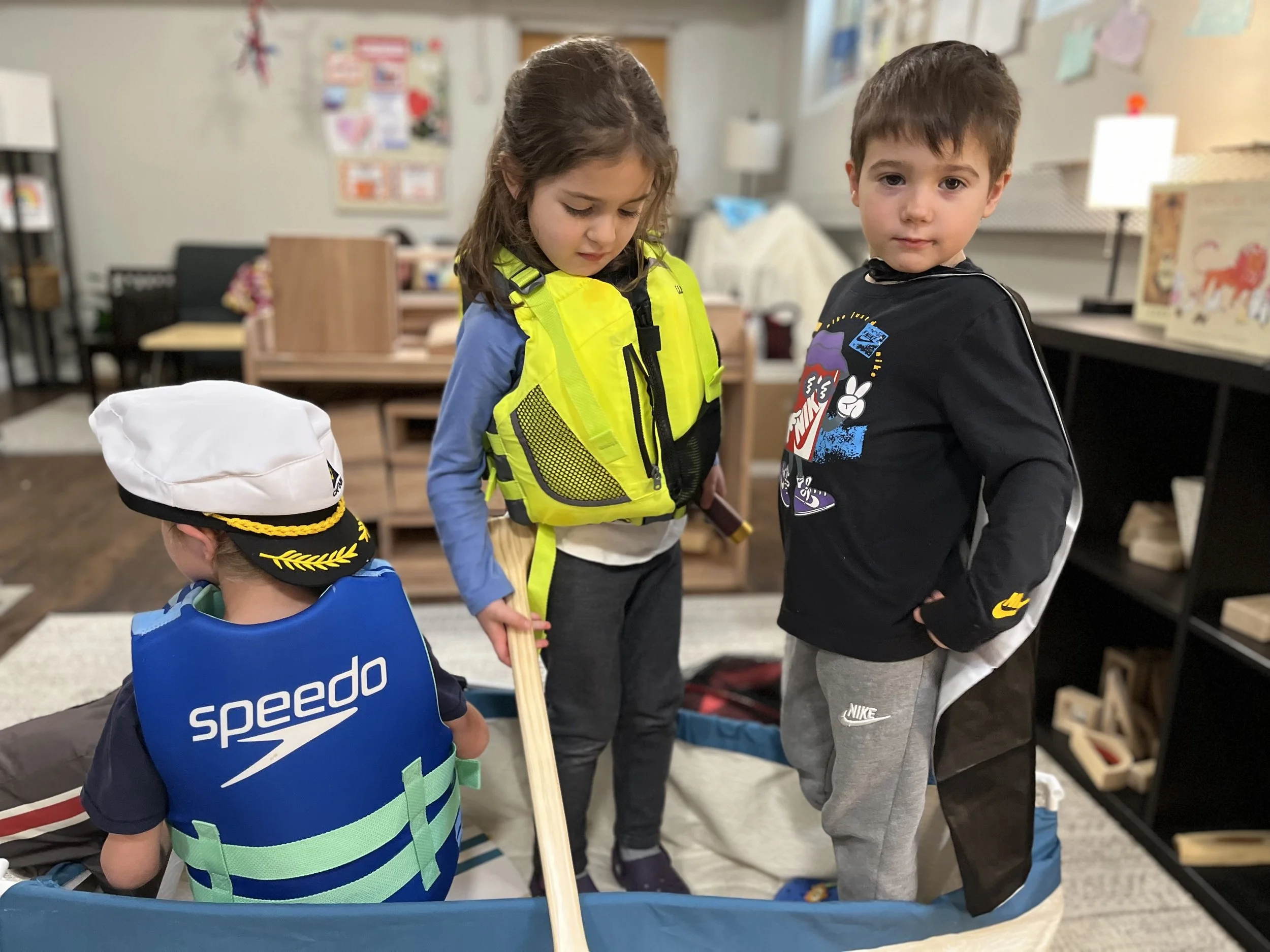 Three young children in costume playing in a classroom, one dressed as a captain, one with a detective or explorer outfit, and one as a ninja, engaging in pretend play.